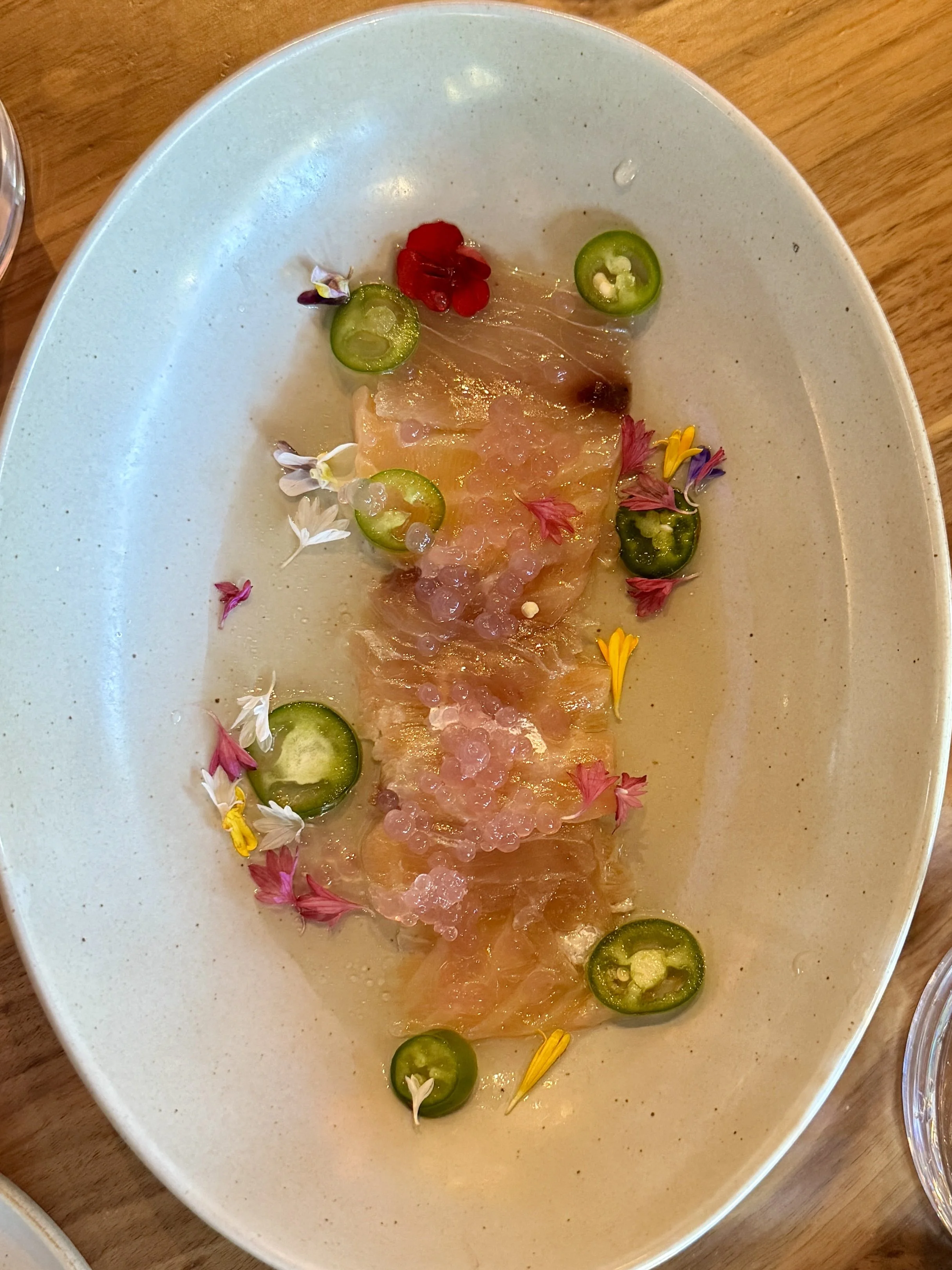 A white oval plate with thin slices of raw fish topped with pinkish fish roe, garnished with sliced jalapeño peppers, edible flowers, and small edible petals on a wooden table.