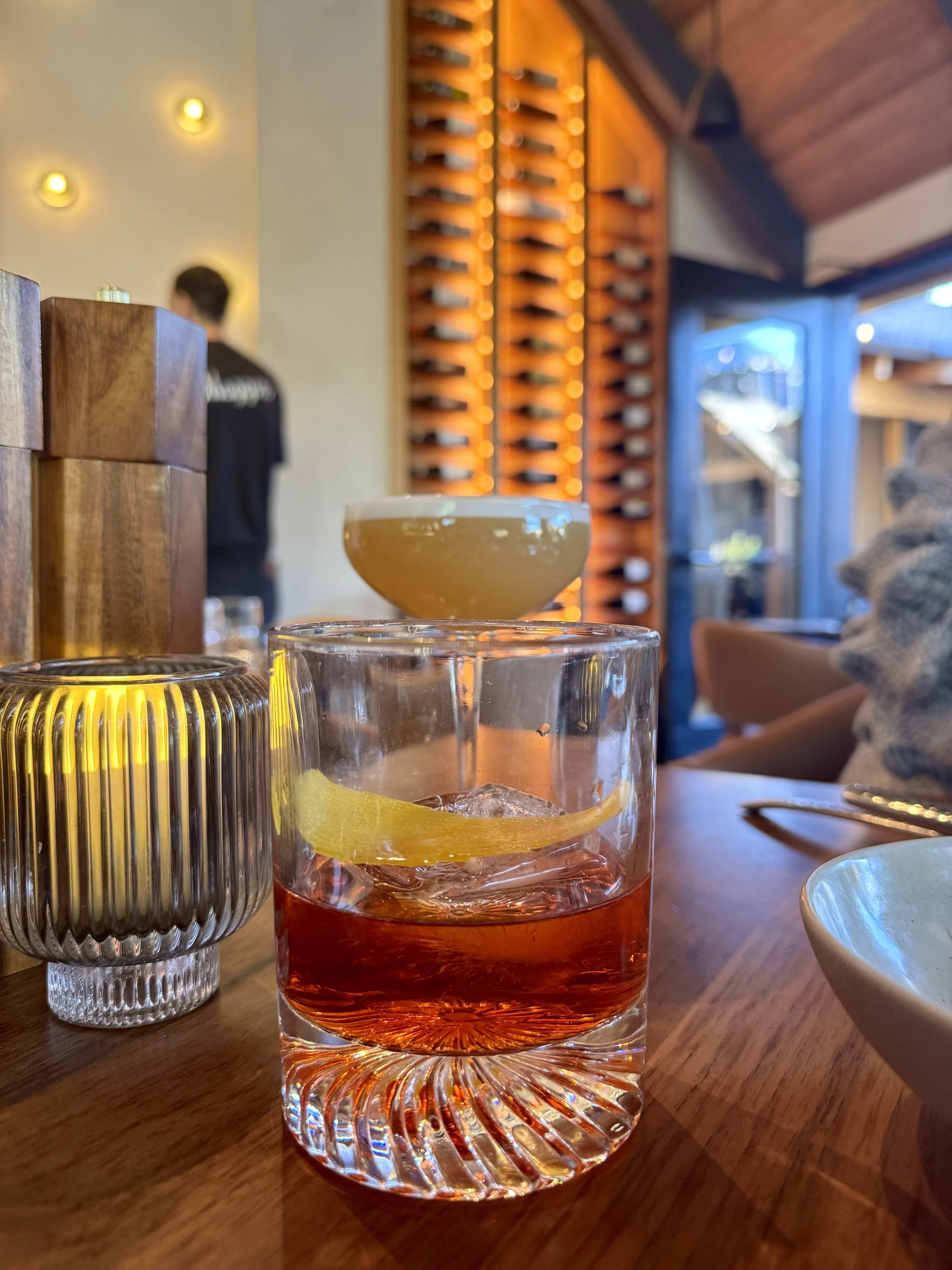 A glass of red orange drink garnished with a lemon slice, placed on a wooden table. There is a metal candle holder, a wooden block, and blurred background of restaurant interior with wine rack and seating.