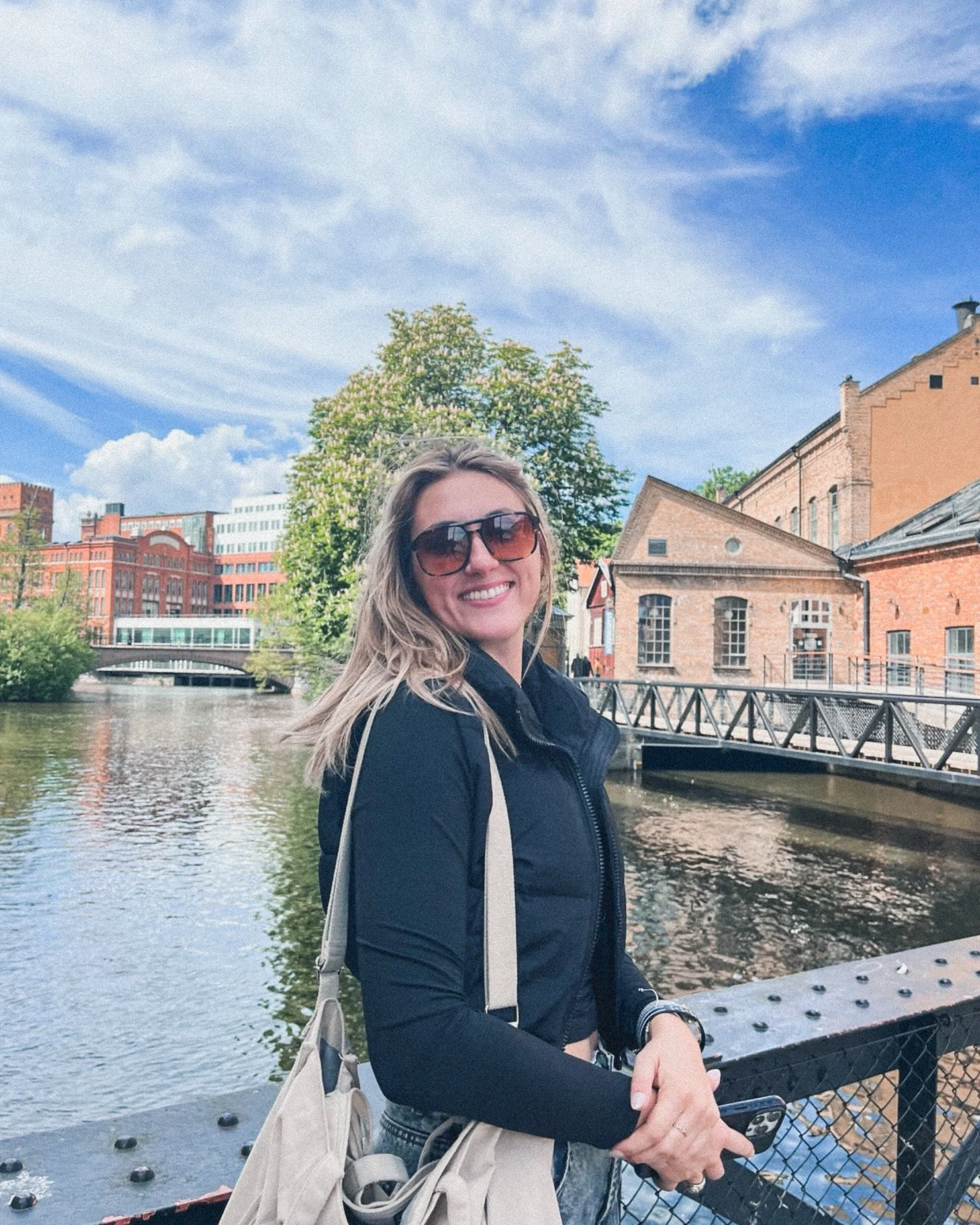 A woman with long blonde hair wearing sunglasses and a black jacket smiling while standing by a river, with historic brick buildings and a bridge in the background, under a partly cloudy sky.