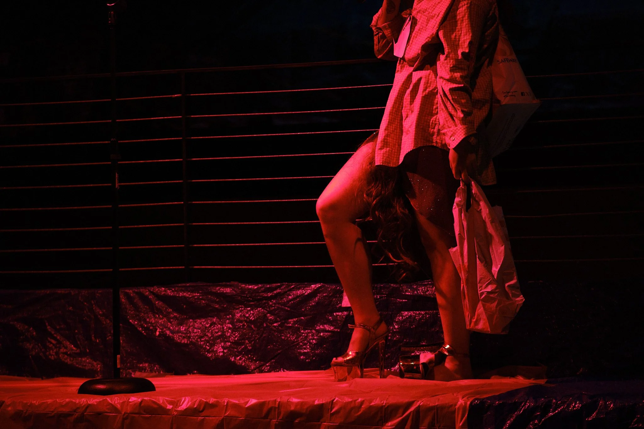 A person in a checkered shirt holding what appears to be a box, standing over a woman with long hair in high heels, on a stage with red lighting and a black background.