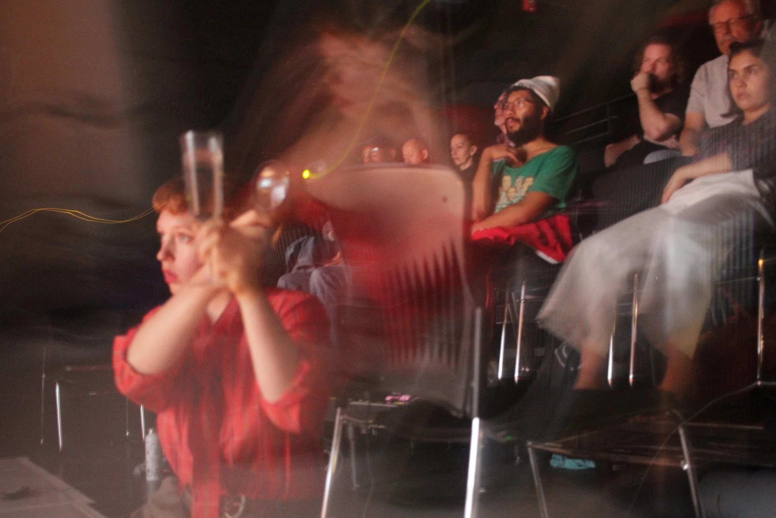 People sitting at tables in a darkened indoor space, possibly a theater or cinema, watching a performance or movie. The background is blurry with some people paying attention to something off-camera.