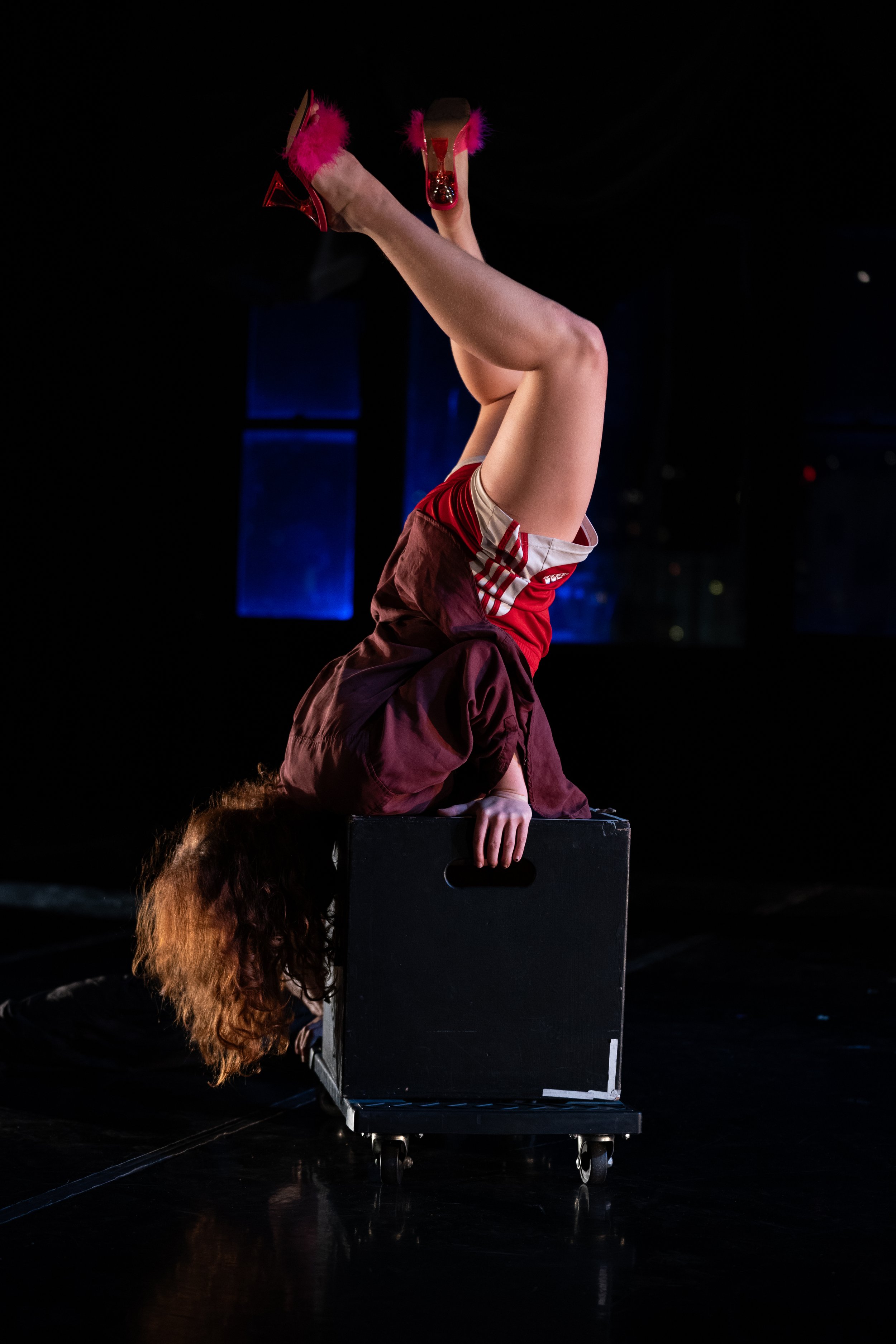 A performer balancing on their hands with legs extended upwards, wearing red shoes with pink feathers and a red and beige outfit, on a black box against a dark background.