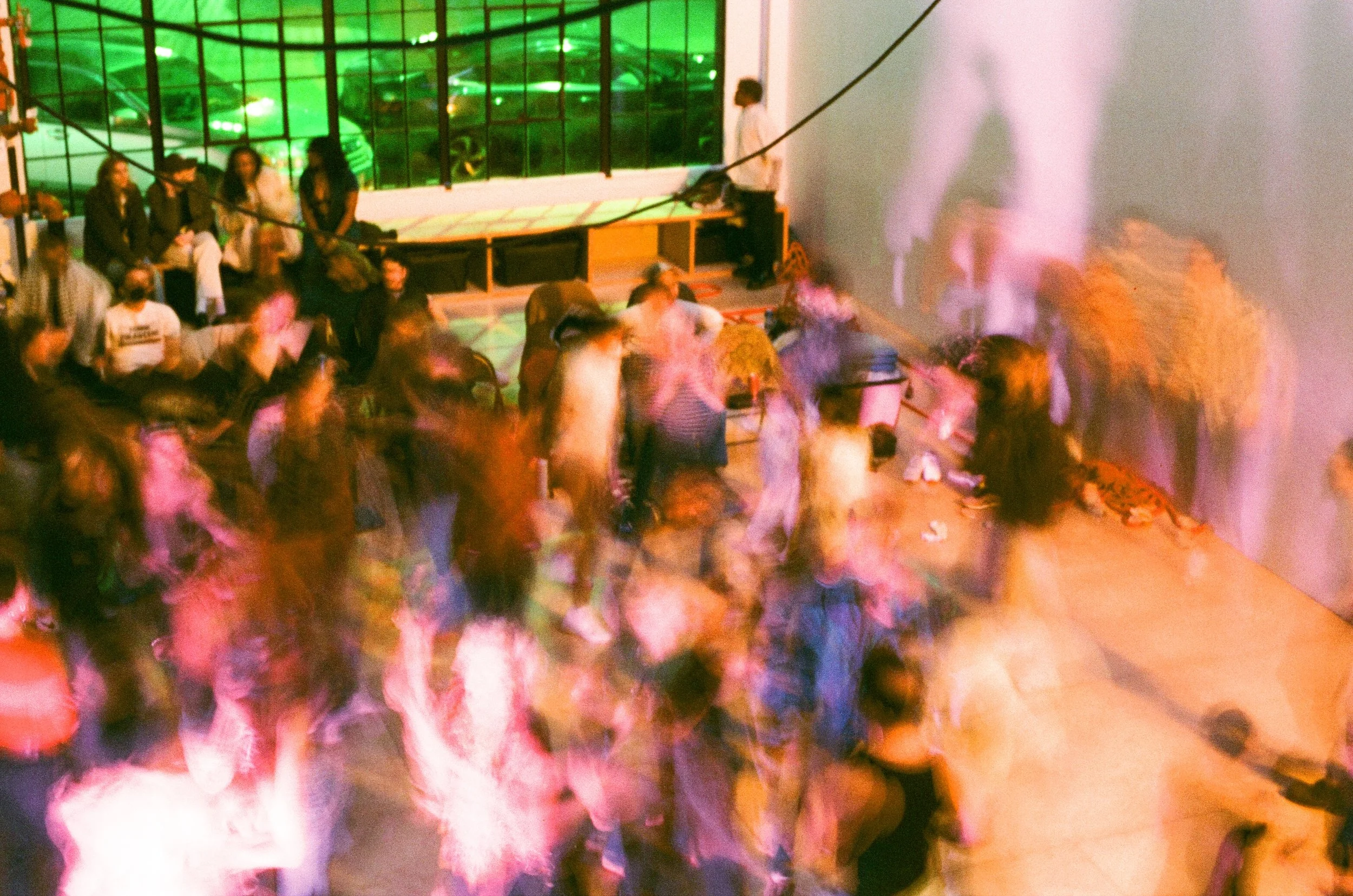 A dimly lit indoor event with a group of people dancing and socializing. There is a large window with green reflections, and a man standing against the wall in the background.