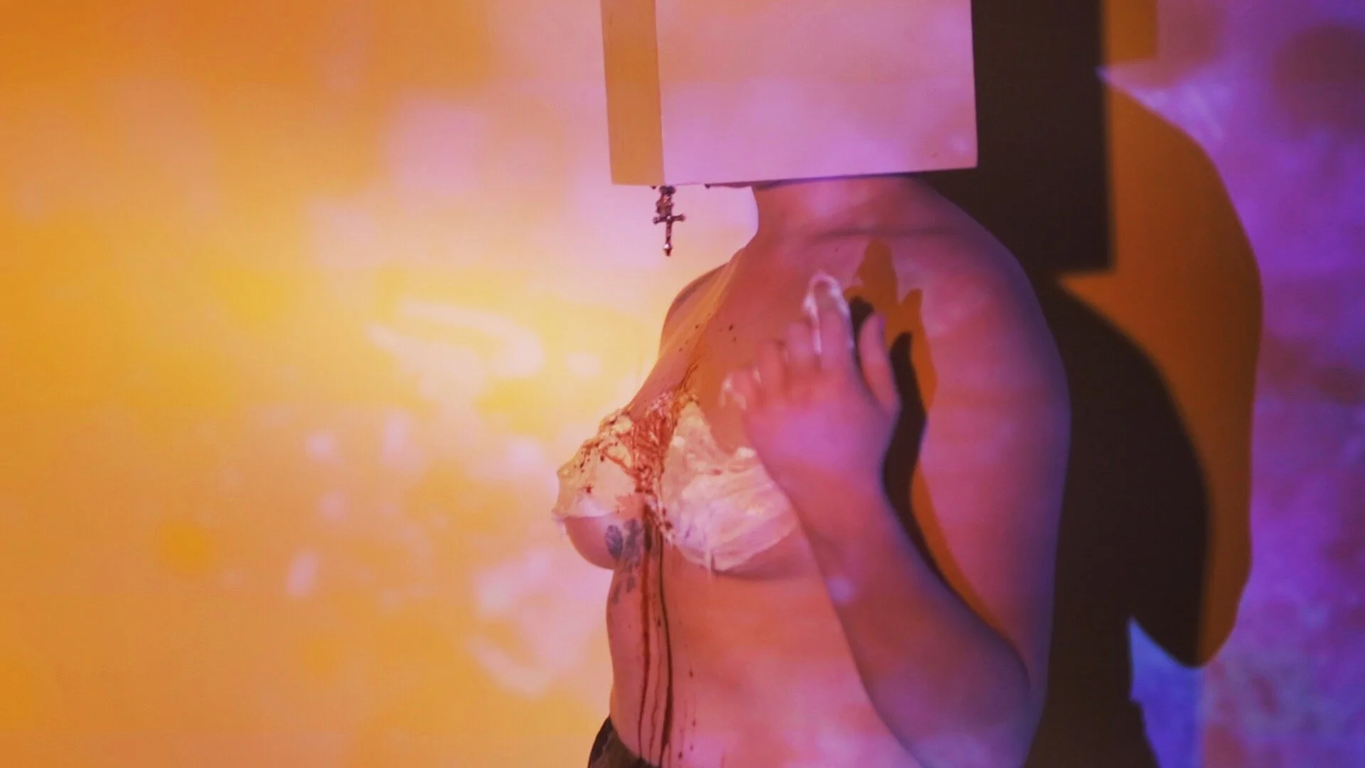 A person with tattoos on their arm leaning over a surface, with colorful purple and yellow lighting, and a white box or panel above their shoulder with a small cross hanging from it.