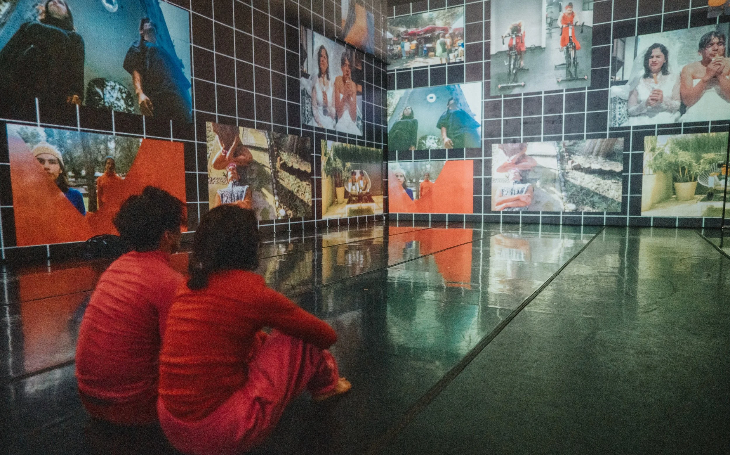 Two children sitting on the floor watching an art exhibition with photographs displayed on black grid walls.
