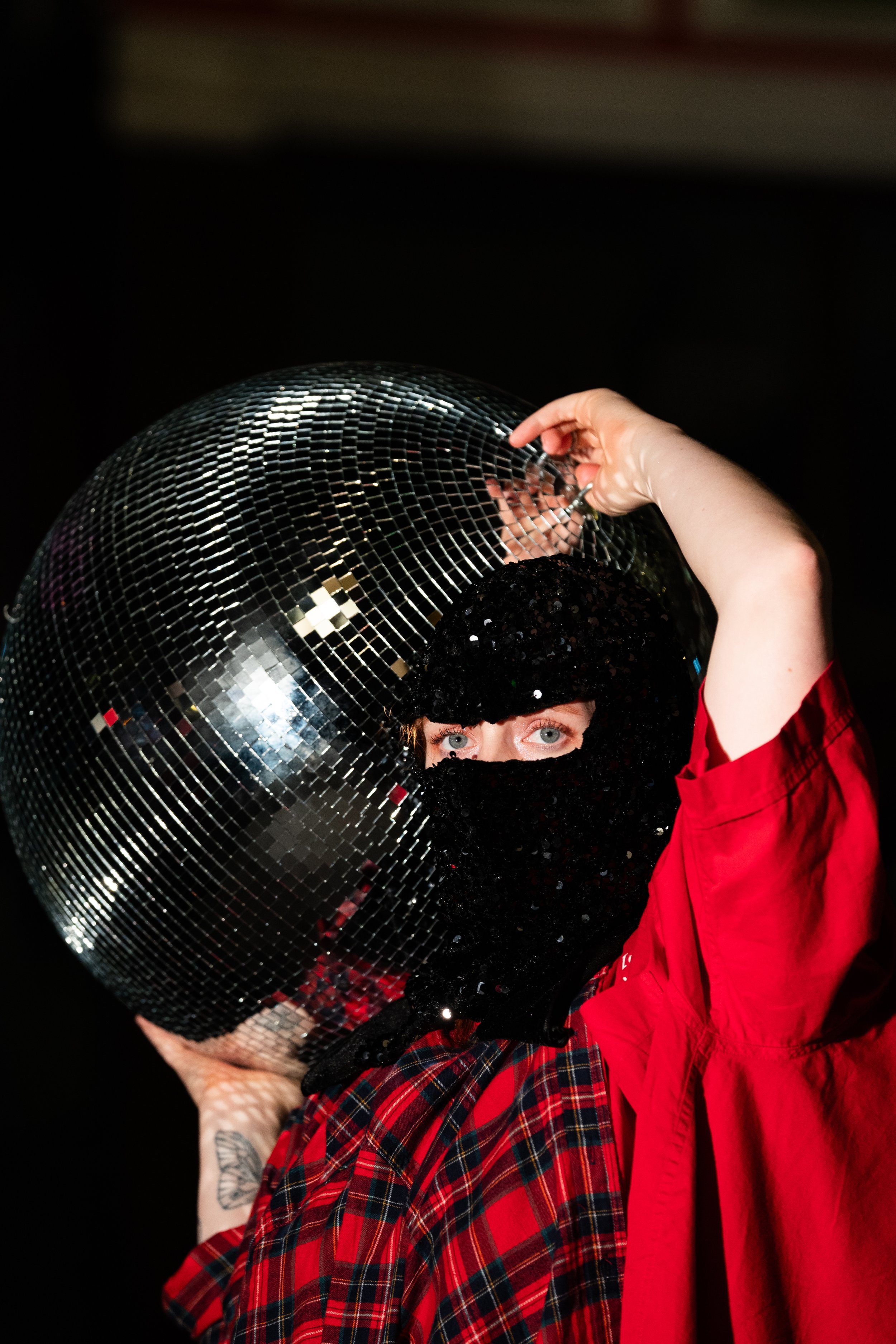 Person with blue eyes holding a large disco ball, wearing a black sequined hat and a red jacket, against a dark background.