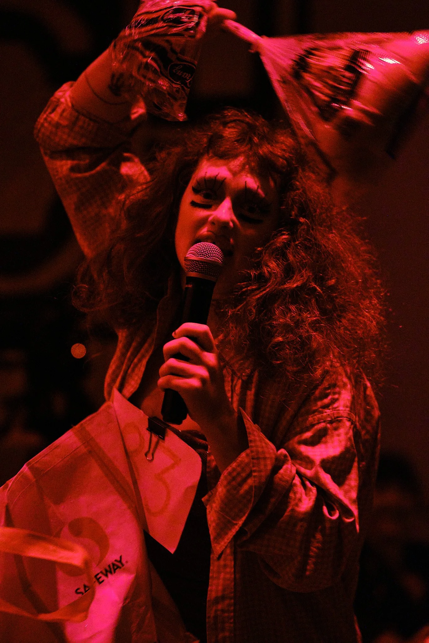 A person with curly hair singing into a microphone, wearing makeup with drawn eyelashes and lines on their face, and holding a plastic bag. The scene is illuminated with red light, giving a dramatic, intense atmosphere.