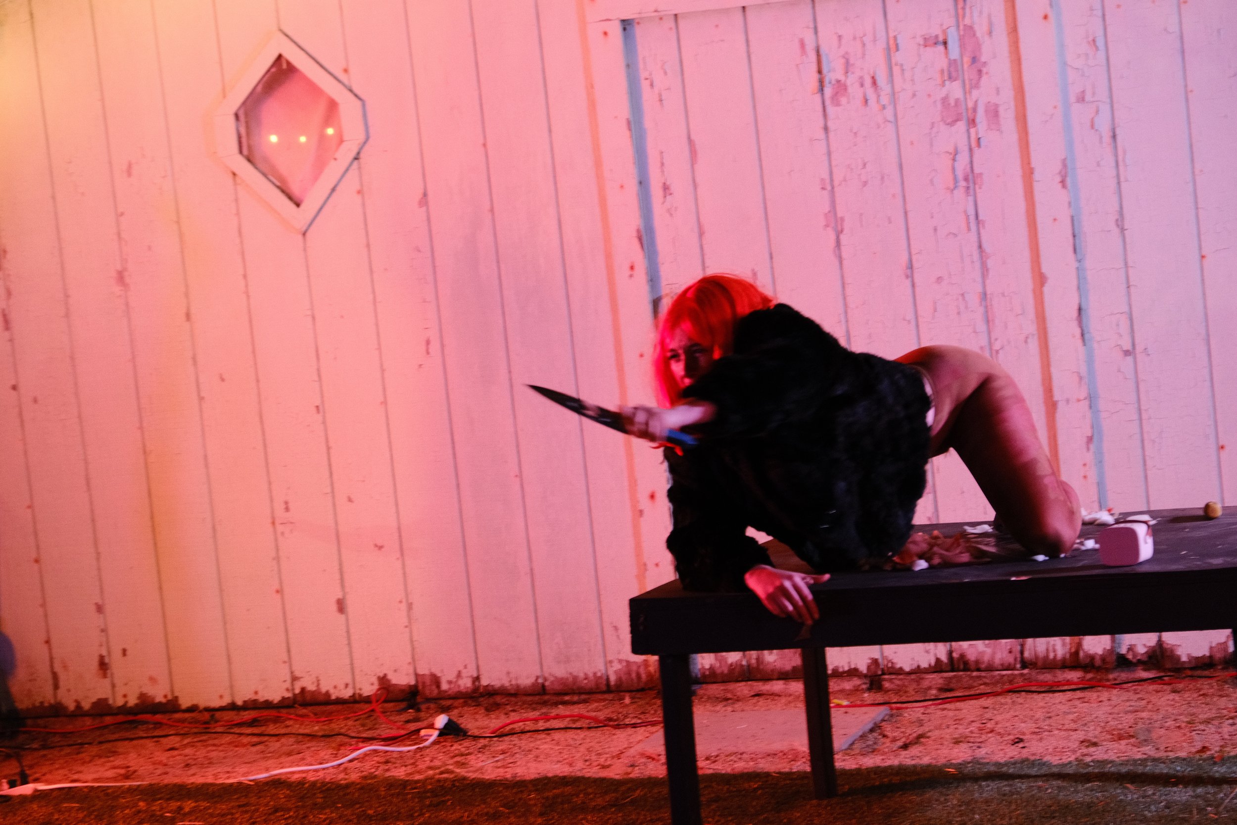 A woman with bright pink hair lying on a stage floor, holding a knife, with a man in black behind her. The stage has a rustic wooden wall with peeling paint and a hexagonal window, illuminated by red lighting.