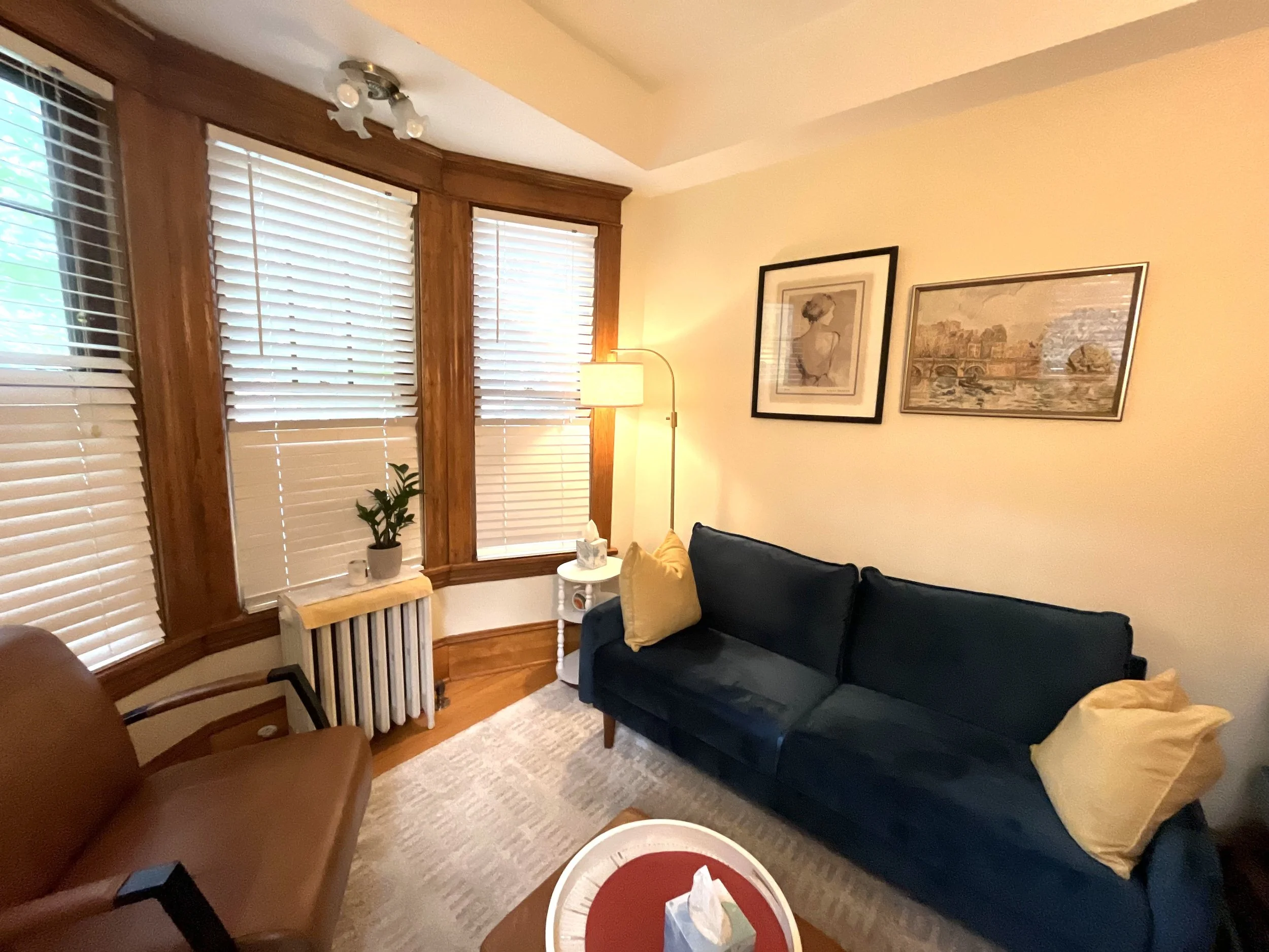 A cozy living room with large window blinds, a brown armchair, a blue sofa with yellow pillows, a small white side table with a plant, framed artwork on the wall, a floor lamp, and a radiator.