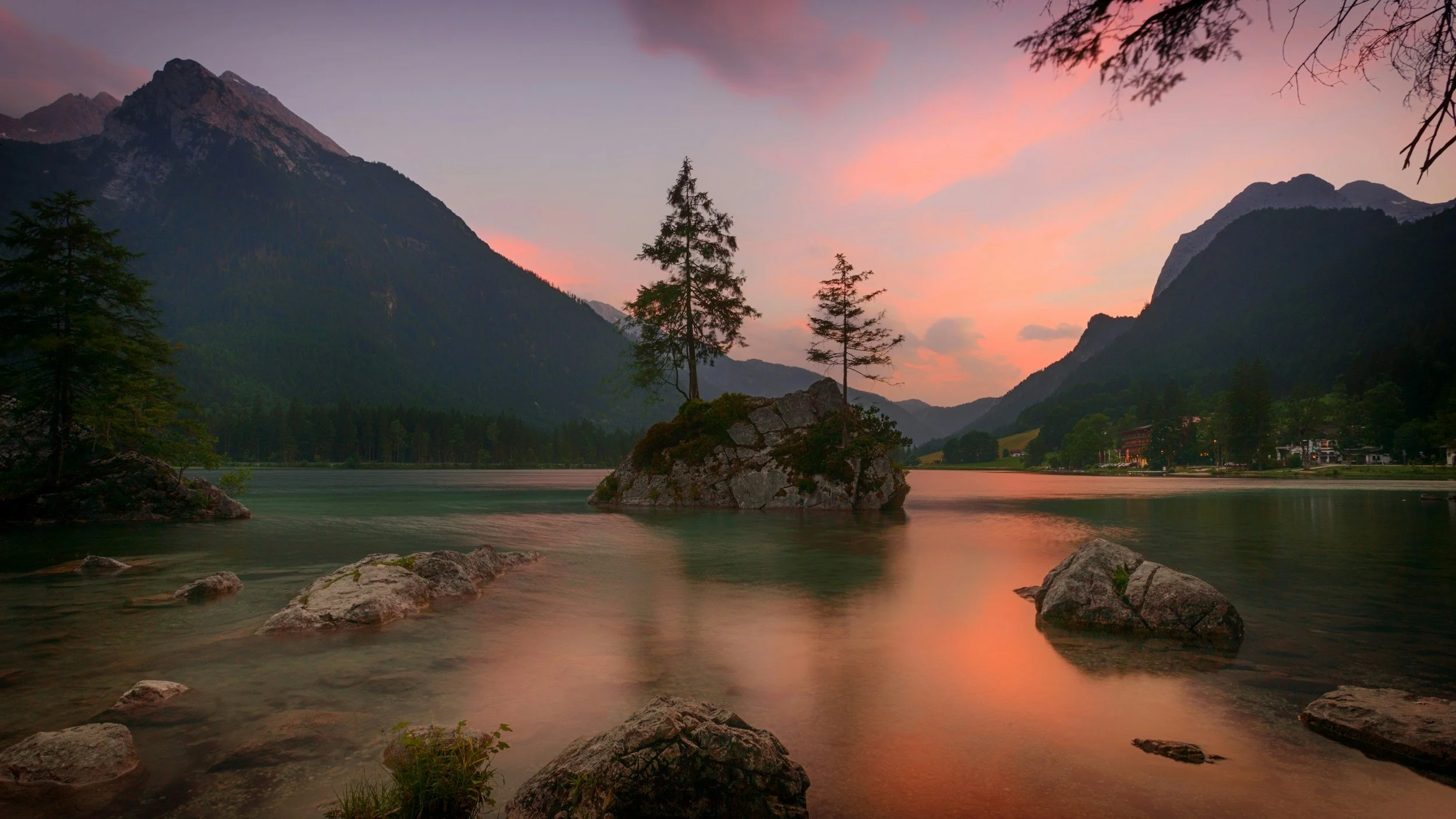 Scenic view of a lake with calm water, surrounded by mountains and trees at sunset, with pink and orange sky.