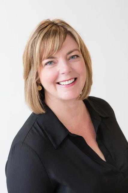 A woman with shoulder-length light brown hair, blue eyes, and a big smile, wearing a black blouse against a light gray background.