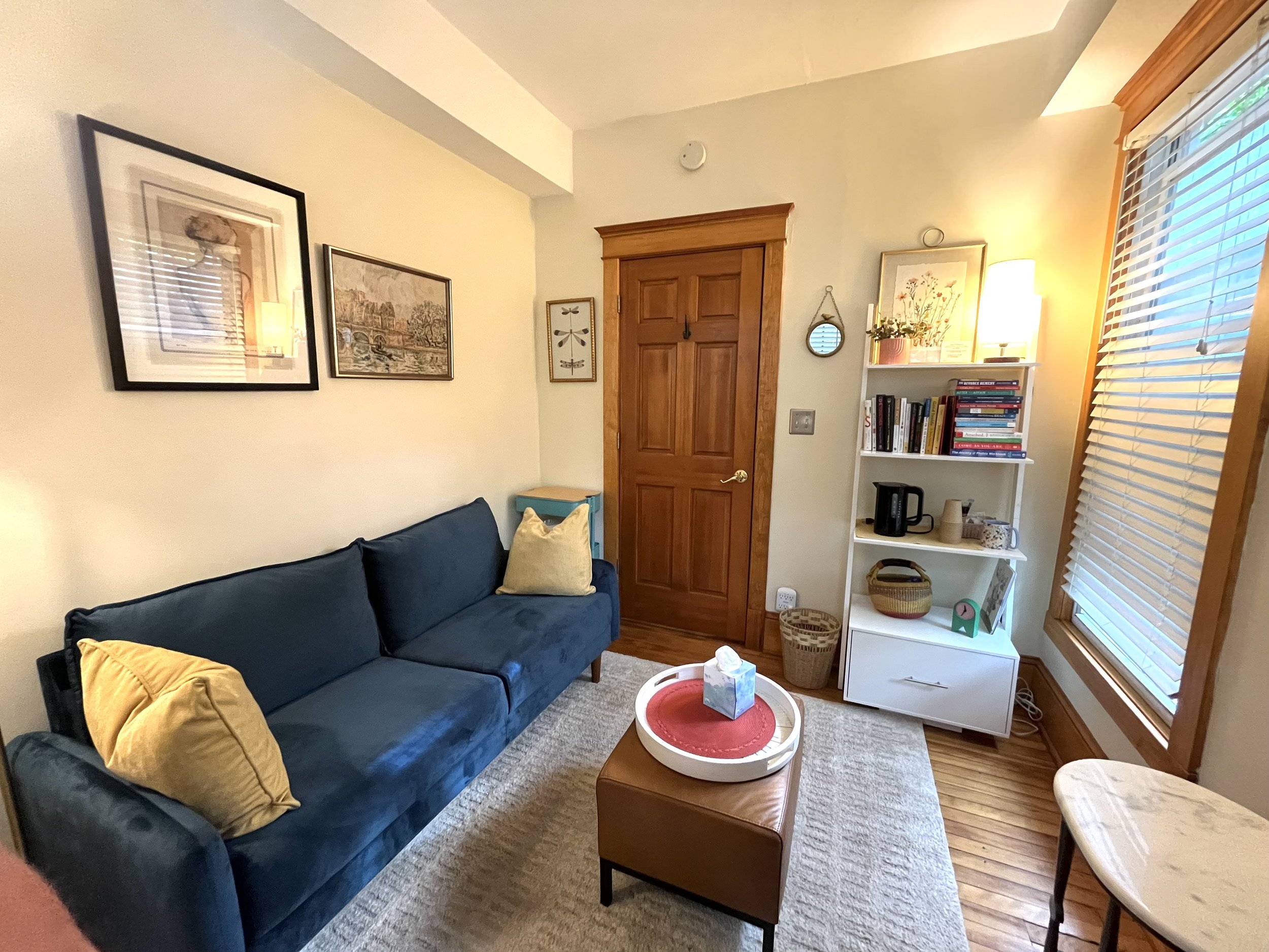A cozy living room with a navy blue sofa, yellow pillows, and a wooden coffee table with a tissue box. There are framed pictures and art on the cream-colored walls, a wooden door, a white bookshelf with books and decorative items near a window with blinds, and a small round mirror hanging on the wall. A lamp on the bookshelf emits warm light, and the room has a carpeted floor.