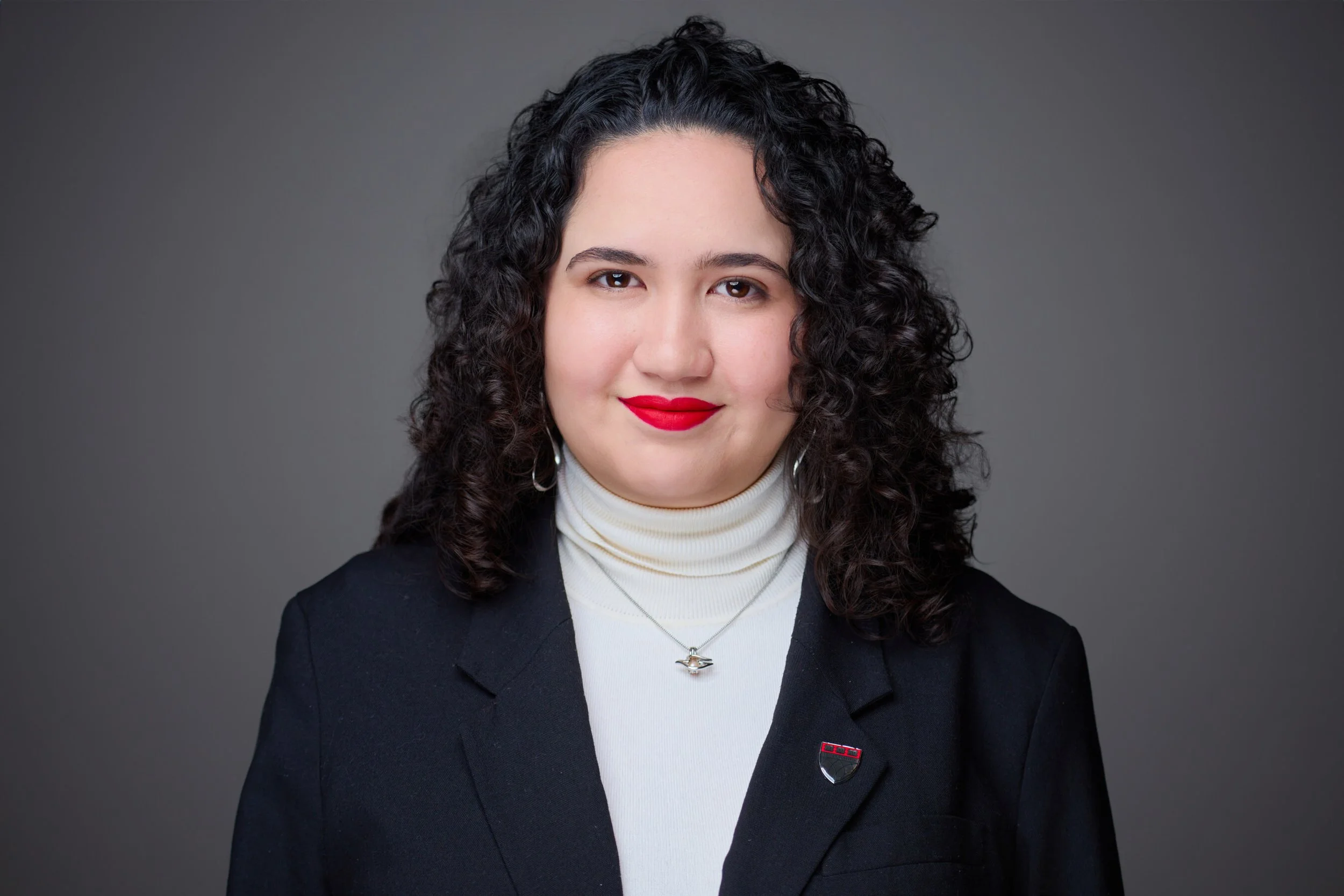 Retrato de la Maestra isabella Leal Aguilar, es una mujer con cabello rizado oscuro, ropa formal negra, labial rojo y aros plateados, frente a fondo gris.