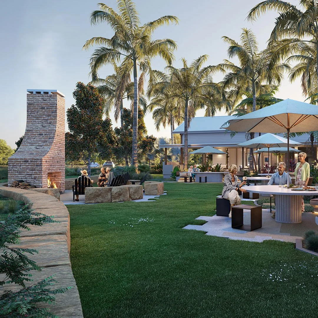 A warm outdoor gathering with multiple groups of people near a brick outdoor fireplace with a fire, in a landscaped yard with lush green grass, tall palm trees, and a modern building with shaded tables and umbrellas.