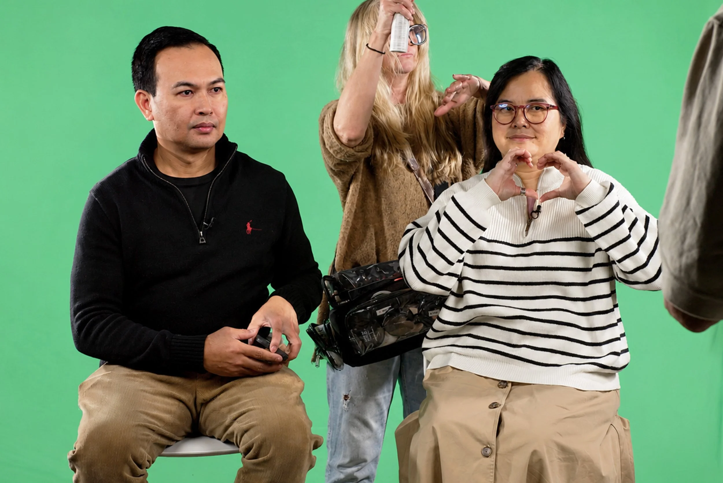 Group of three people against a green screen, two seated men and one standing woman, with a camera and equipment, making hand gestures and posing.