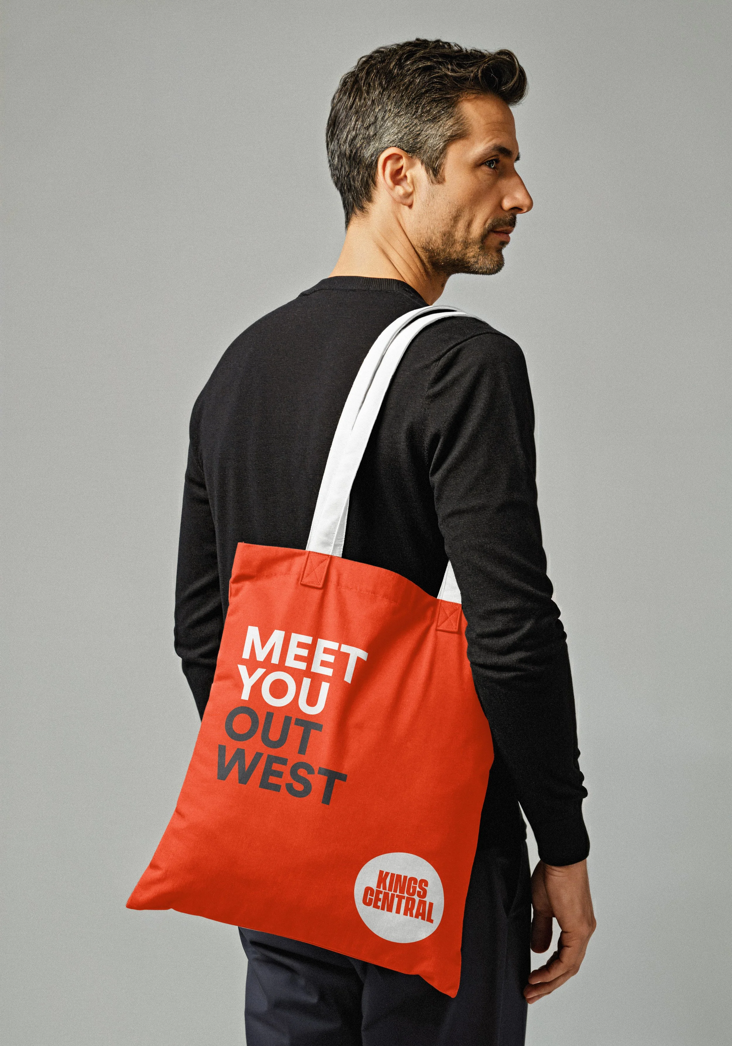 Side view of a man in a black long-sleeve shirt with a red shopping bag over his shoulder that reads "Meet You Out West" and "Kings Central" at the bottom, standing against a plain gray background.