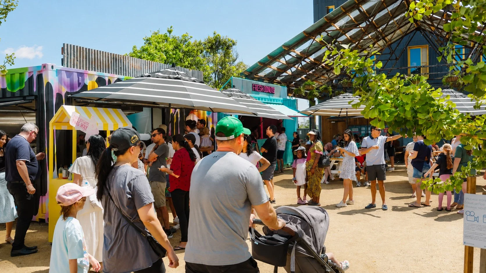 Crowd of people at an outdoor food market with umbrellas, colorful buildings, and trees on a sunny day.