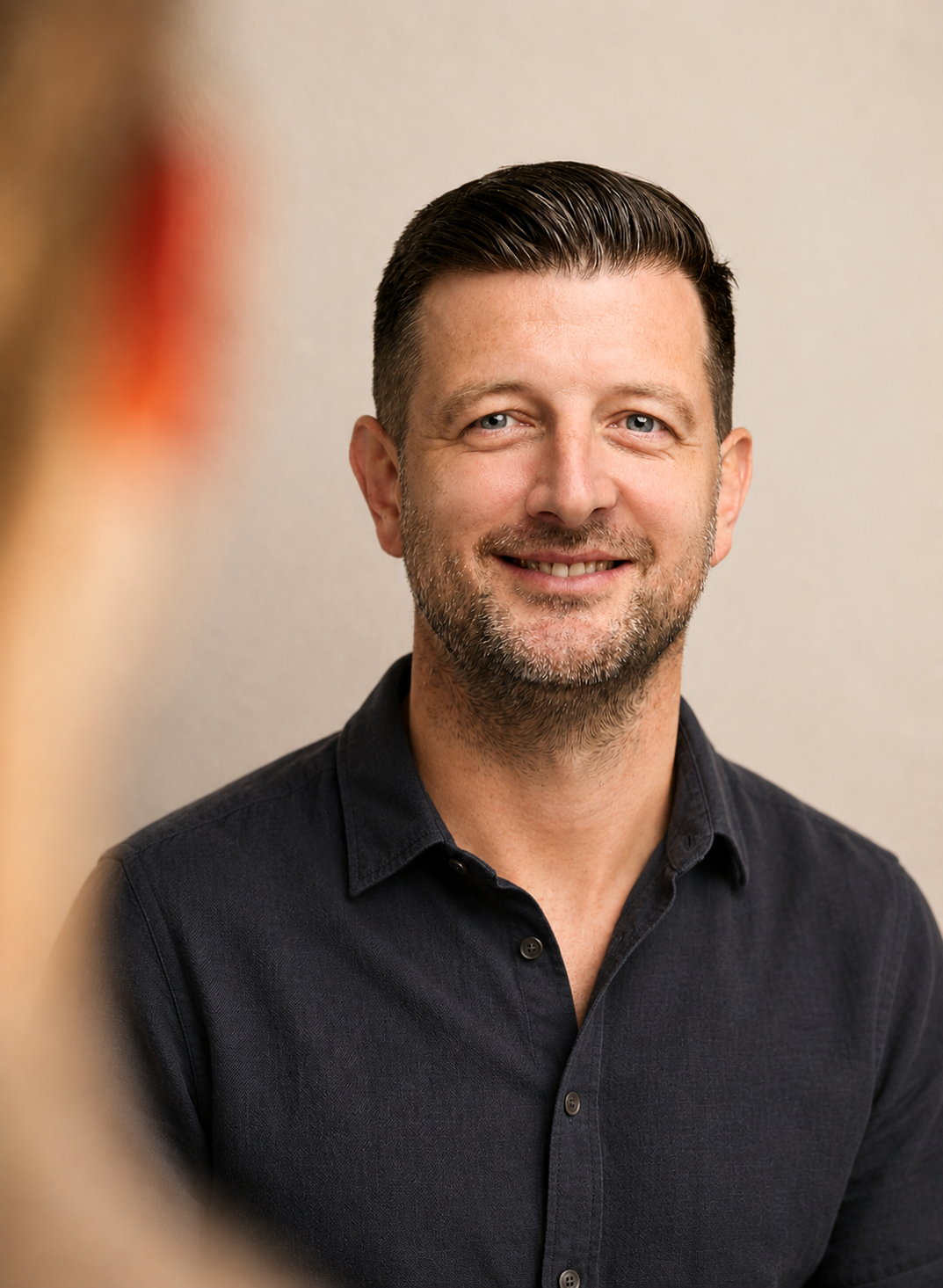 A smiling man with short dark hair and a beard, wearing a black shirt, looking at someone off-camera with a blurred foreground of another person's hair in the left corner.