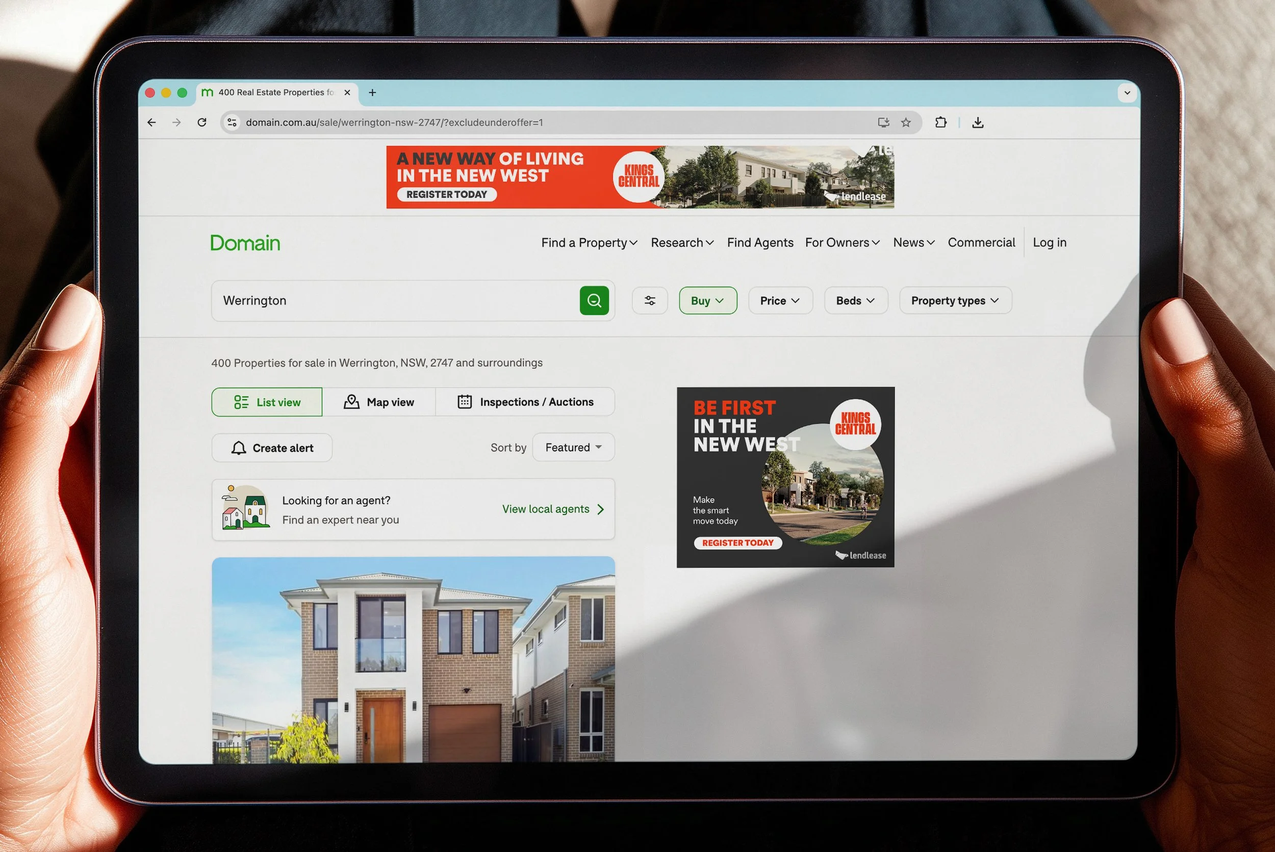 Close-up of a person's hand holding a tablet displaying a real estate website with property listings and advertisements.