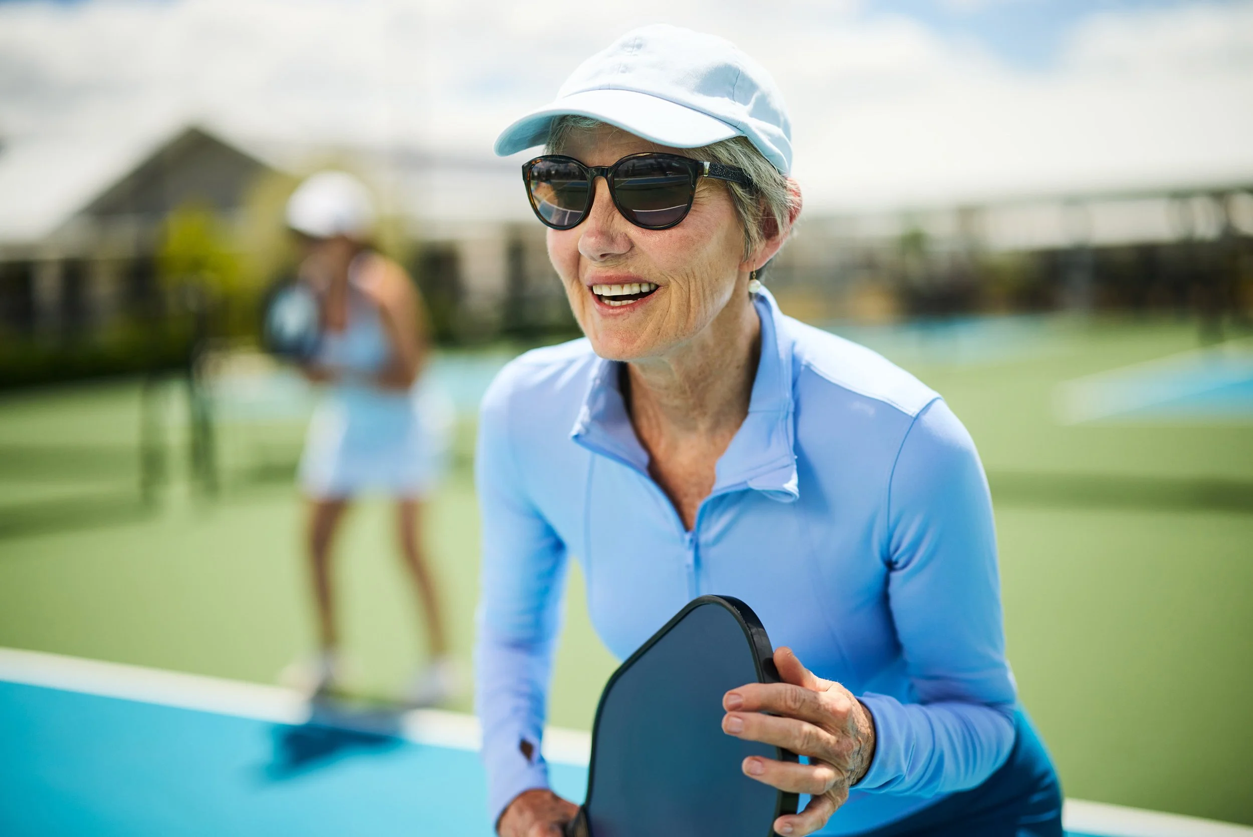Elderly woman smiling, wearing sunglasses, a light blue cap, and a light blue top, holding a paddle tennis racket on an outdoor tennis court.