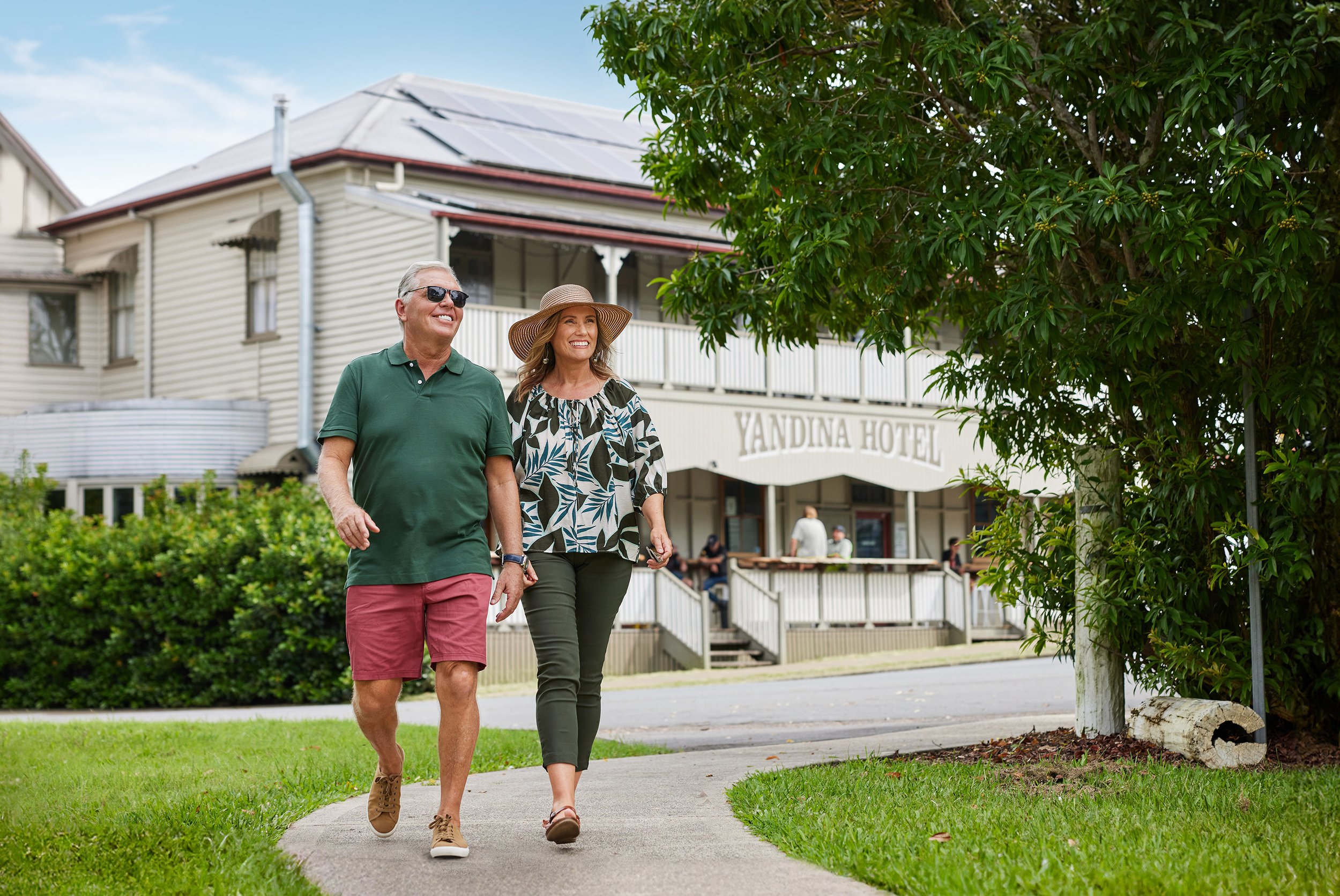 A man wearing sunglasses and a green polo shirt walking with a woman in a hat and patterned blouse on a sidewalk near Yandina Hotel, with greenery and a tree in the background.