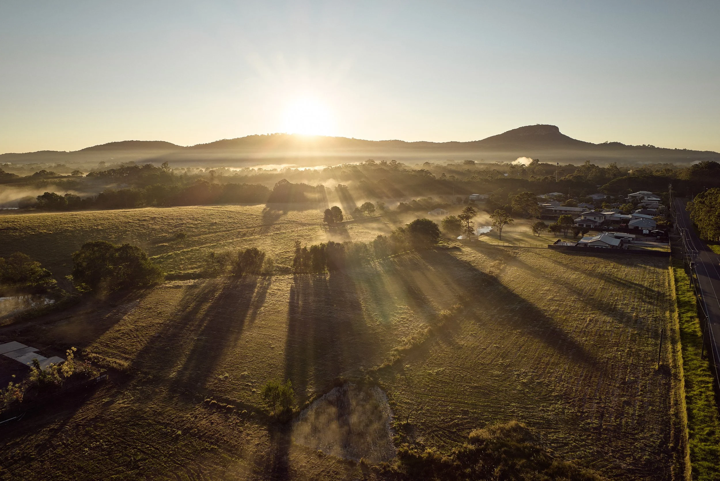 Sun rising over green farmland with trees and a small community, mist in the air, hills in the background, and smoke from chimneys.