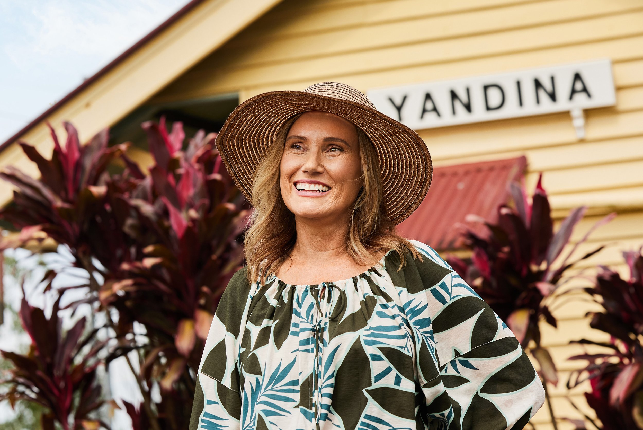 Woman with long wavy hair smiling, wearing a wide-brimmed straw hat and a black and white patterned blouse, standing in front of a yellow wooden building with a sign that reads 'YANDINA' and purple-leafed plants.