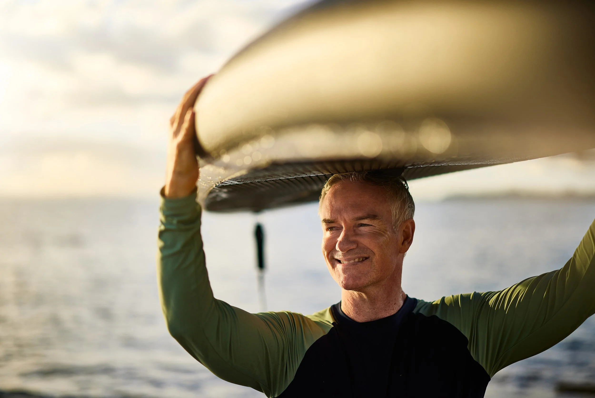 A man smiling while holding a surfboard above his head outdoors near the water during sunset.