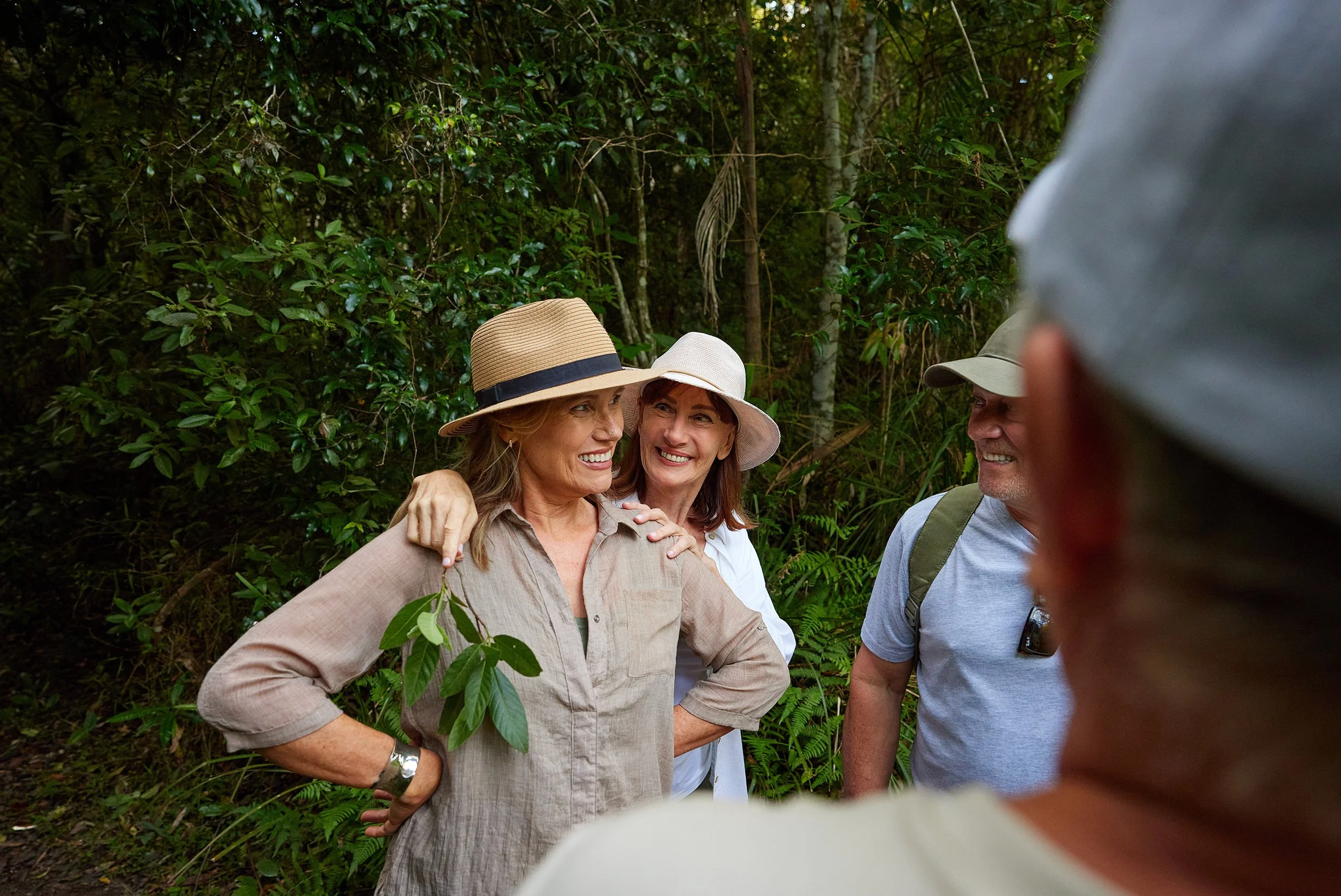 Group of people smiling and enjoying each other's company in a lush green forest, wearing hats and casual outdoor clothing.