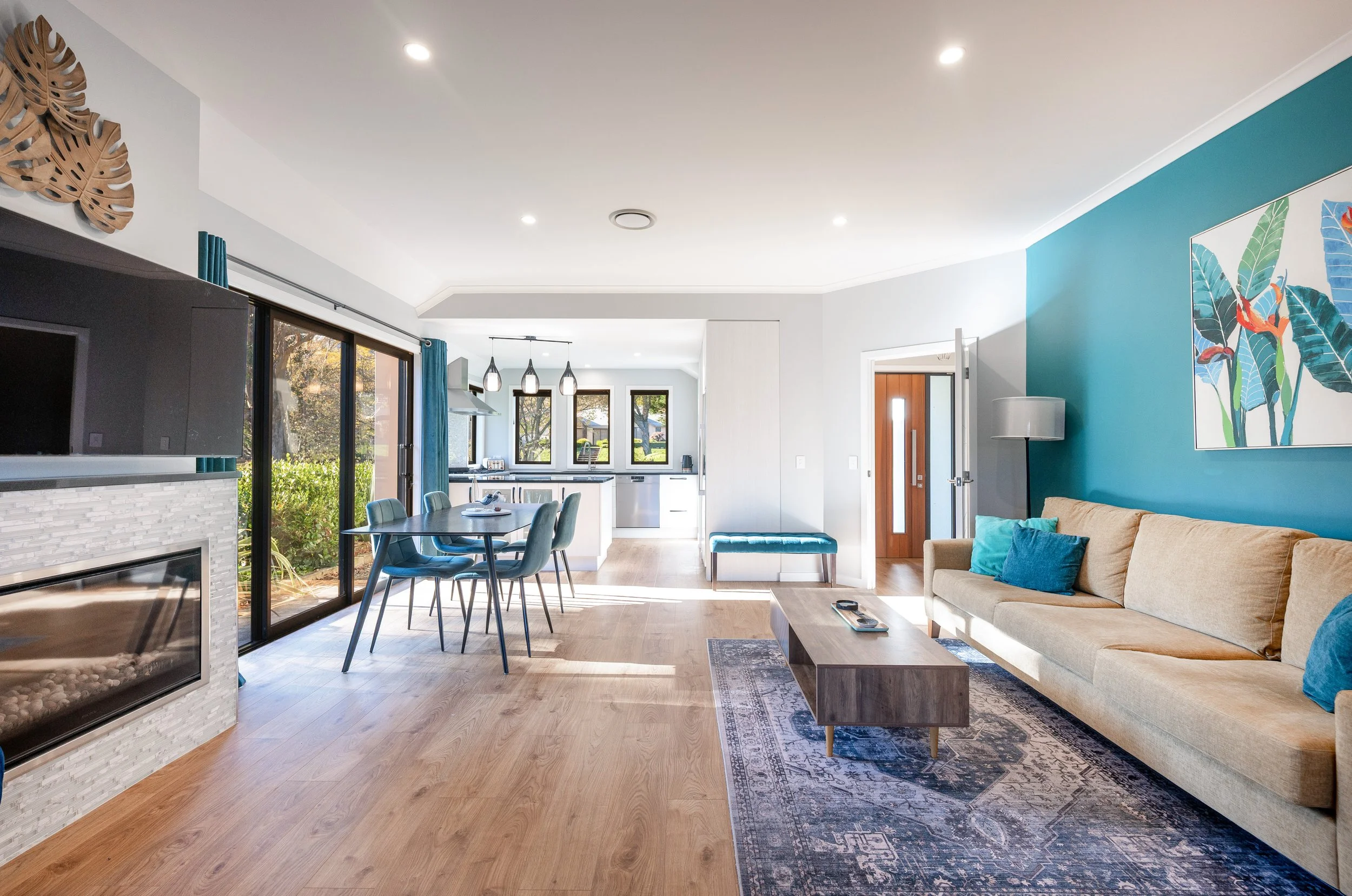 A bright open-concept living and dining area with large windows, a beige sofa with teal and blue pillows, a wooden coffee table, a patterned area rug, a dining table with teal chairs, a white kitchen with pendant lights, and tropical wall art.