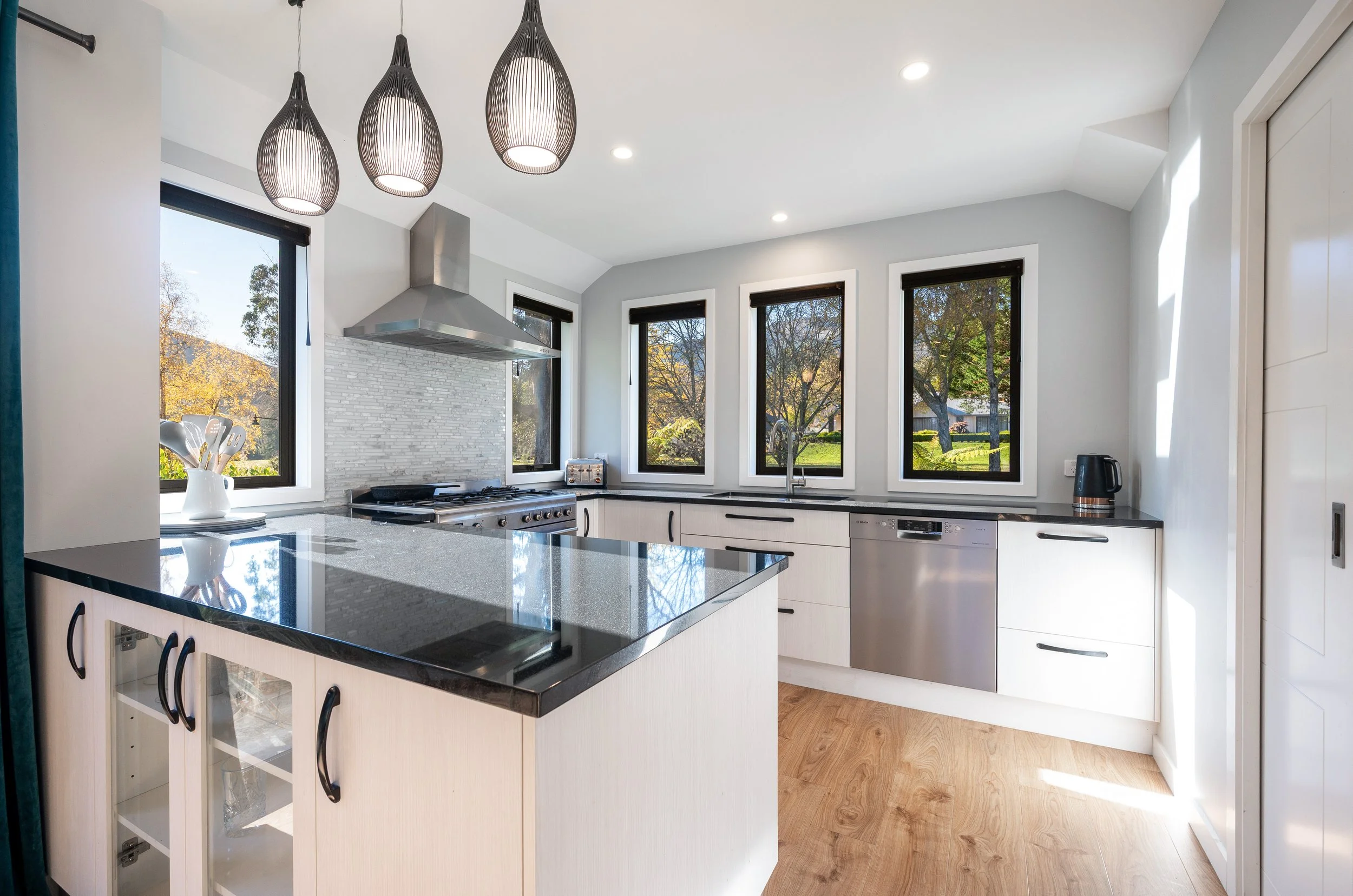 Modern kitchen with white cabinets, black countertops, a stainless steel range hood, and five windows showing trees outside; pendant lights hanging from the ceiling.