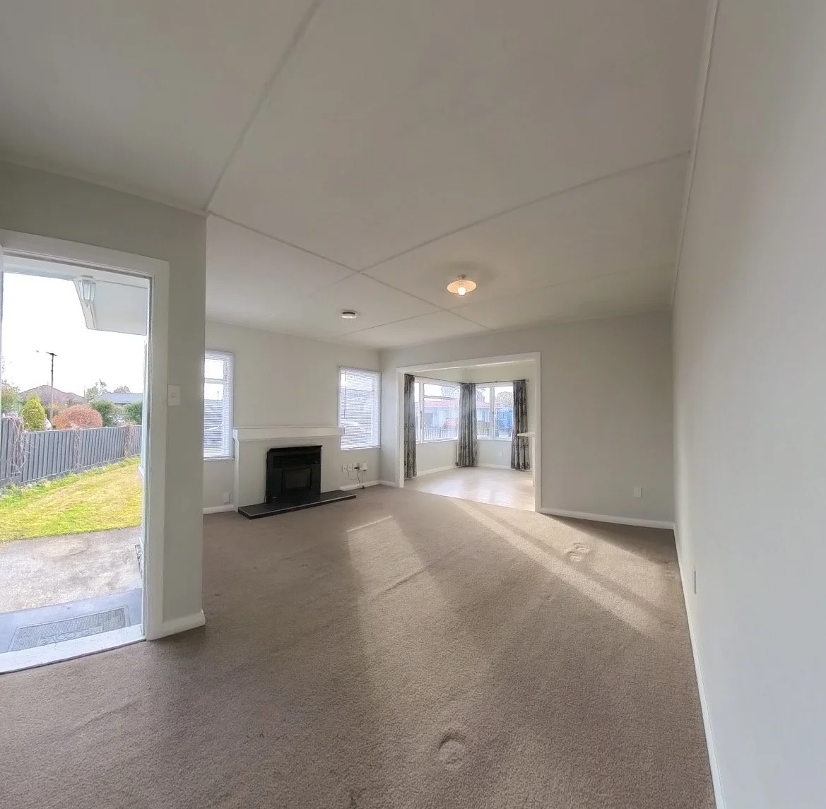 Empty living room with beige carpet, white walls, a fireplace, and windows with curtains, leading to a bright sunroom with large windows and patterned curtains.