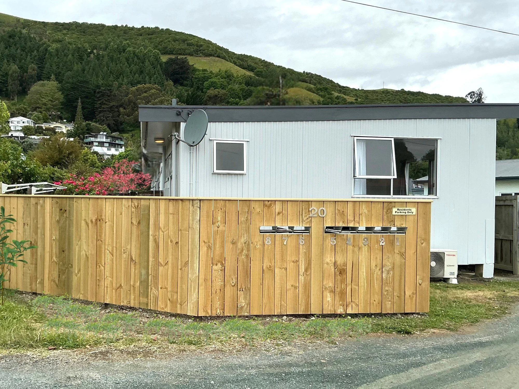 A residential building with a light blue exterior, open windows, and a satellite dish on the roof, surrounded by a wooden fence, with hills and other houses in the background.