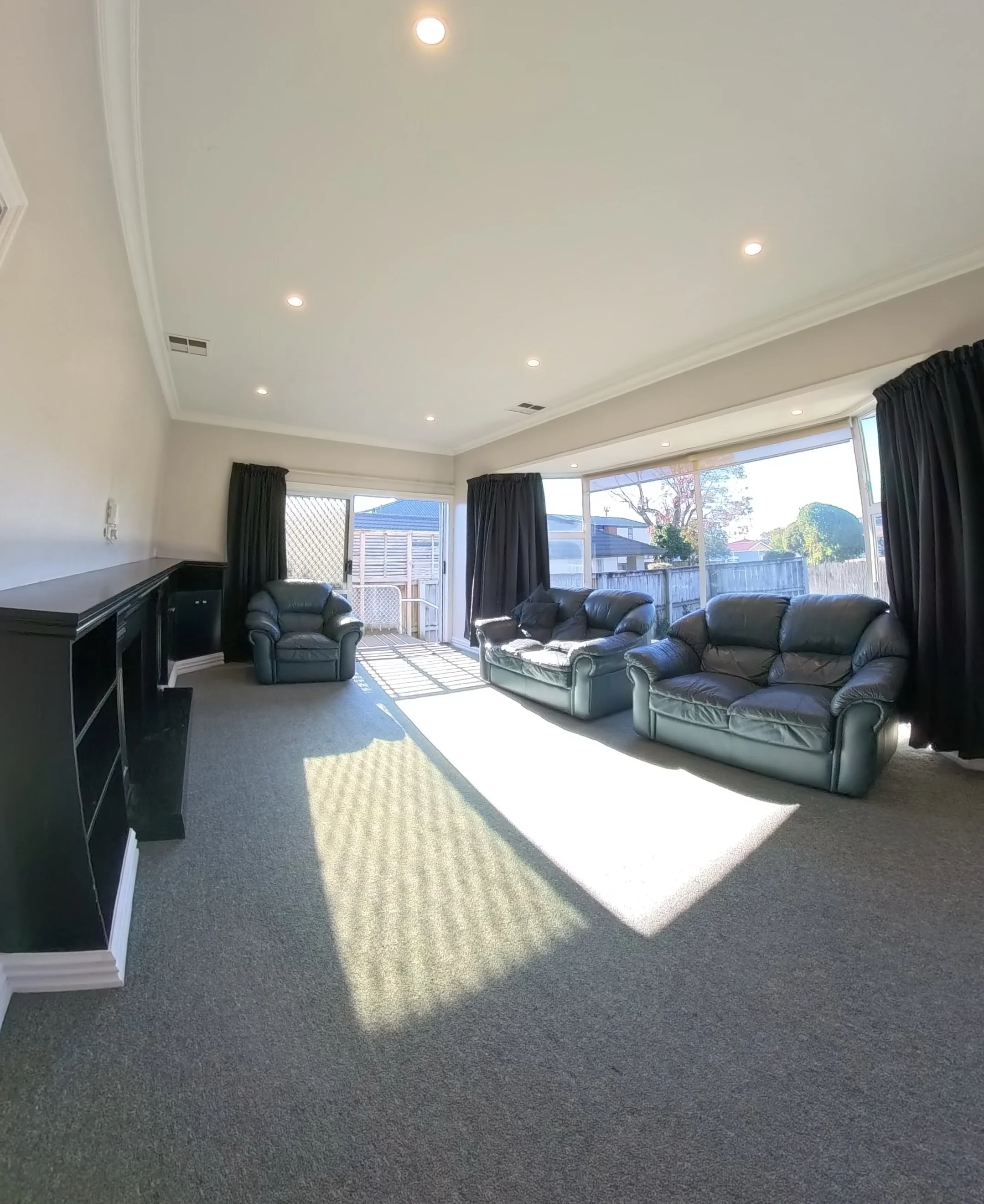 Living room with large windows, blue sofas, black curtains, and gray carpet. Sunlight casts shadows on the carpet.