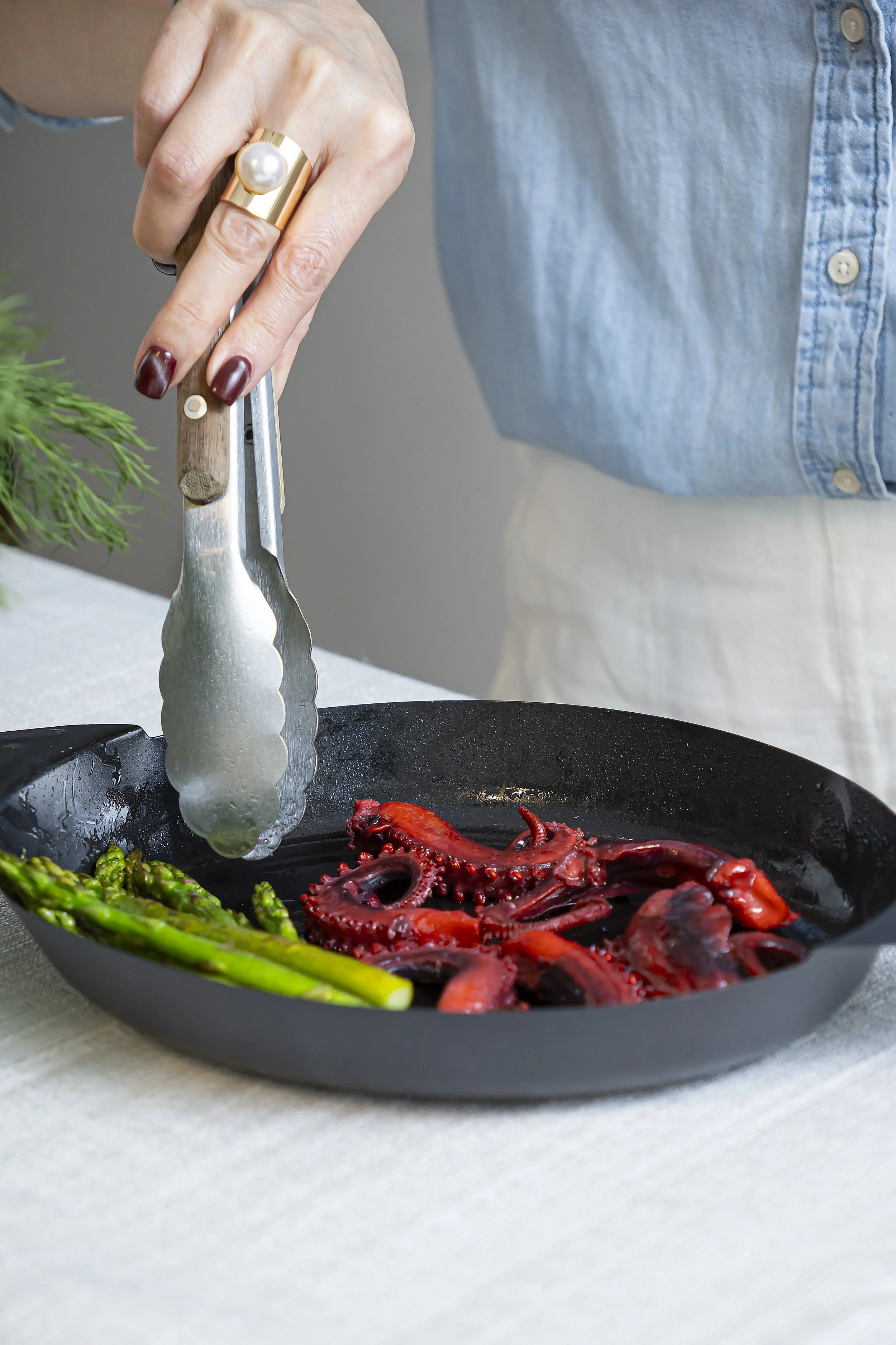Person using tongs to cook red octopus and green asparagus in a black skillet.