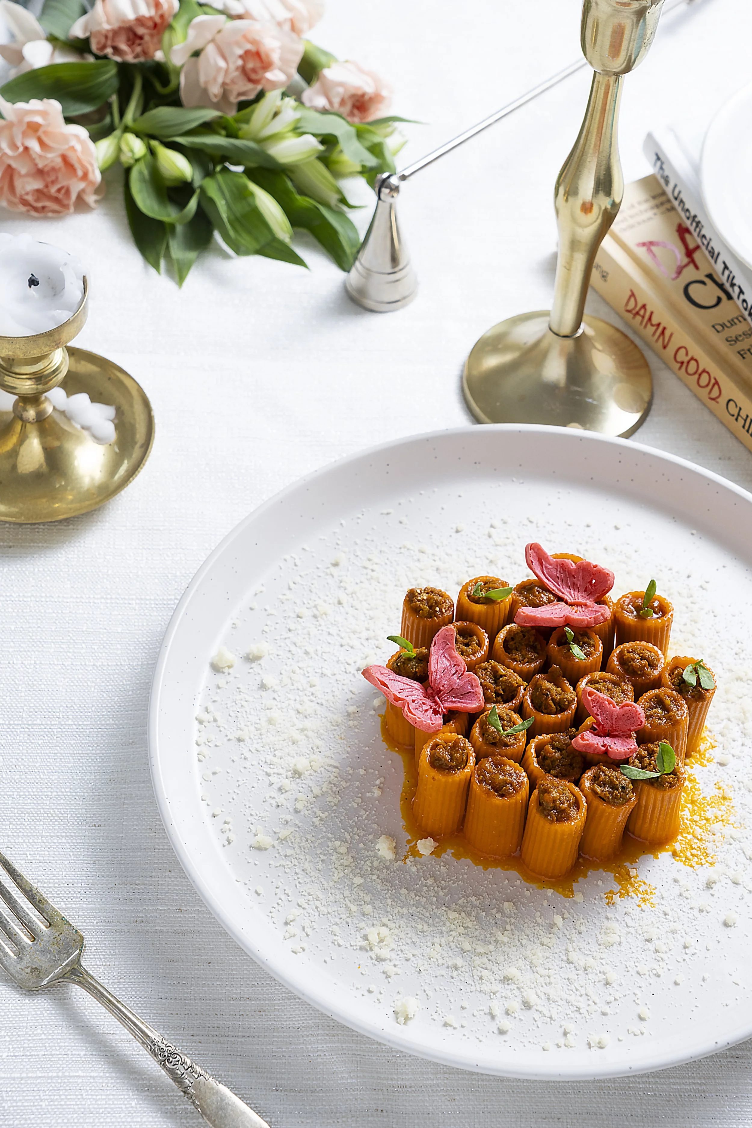 Plate of stuffed pasta with tomato sauce and pink flower garnishes, on a white tablecloth with books and a candle in the background.