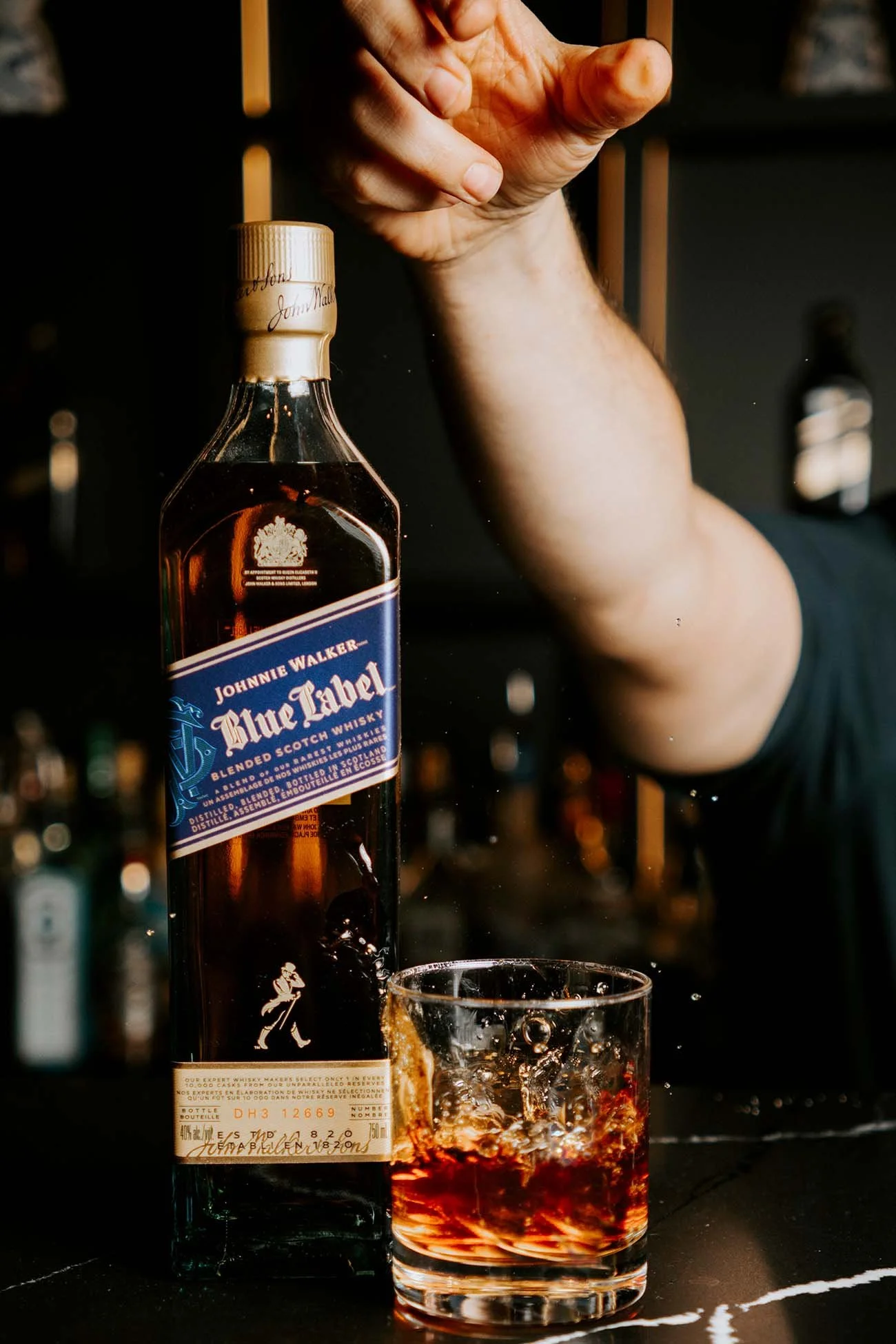 A bottle of Johnnie Walker Blue Label blended Scotch whisky next to a glass filled with ice and dark liquid, with a hand in the background.