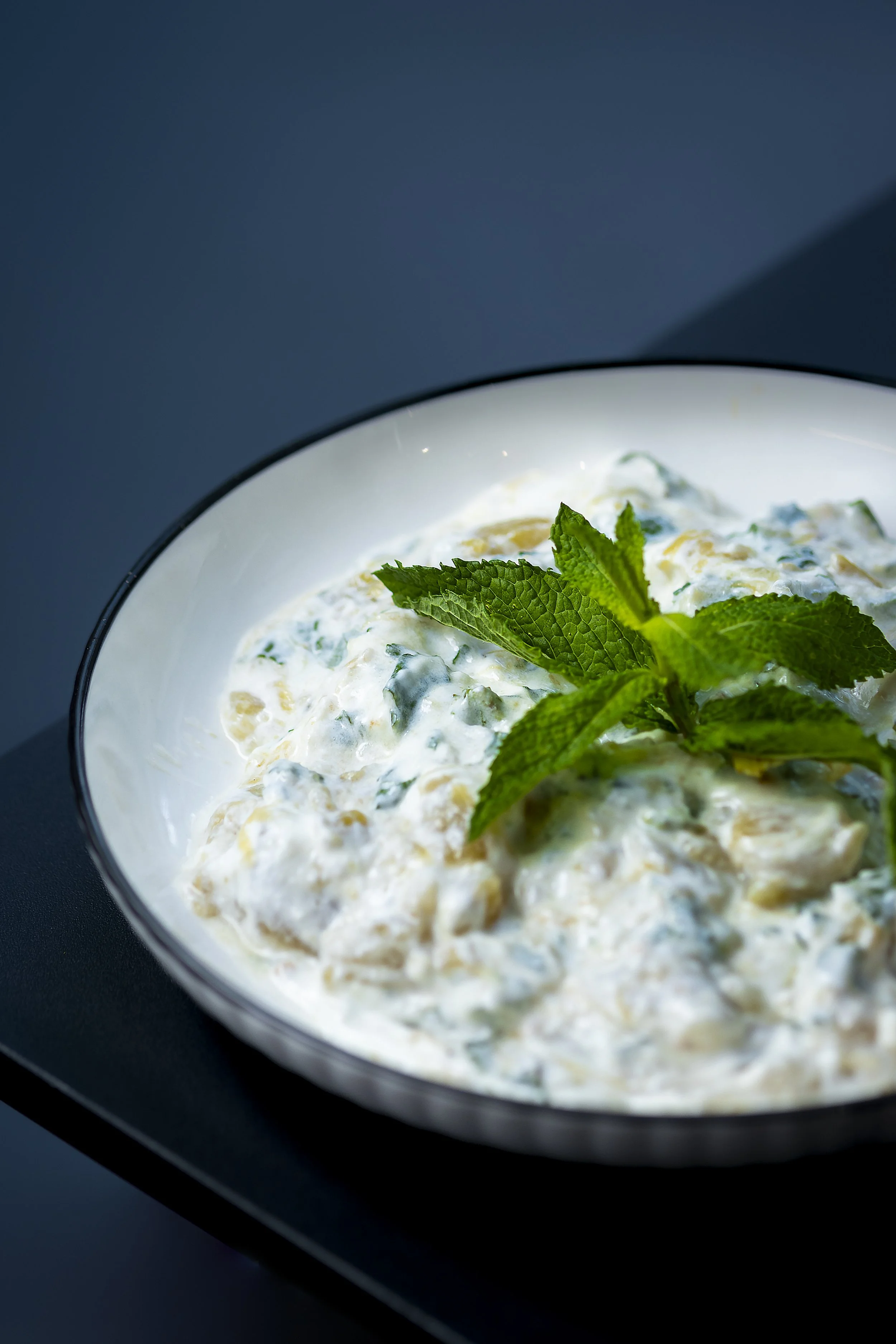 Close-up of a bowl of creamy dip garnished with mint leaves, placed on a dark surface with a plain dark background.
