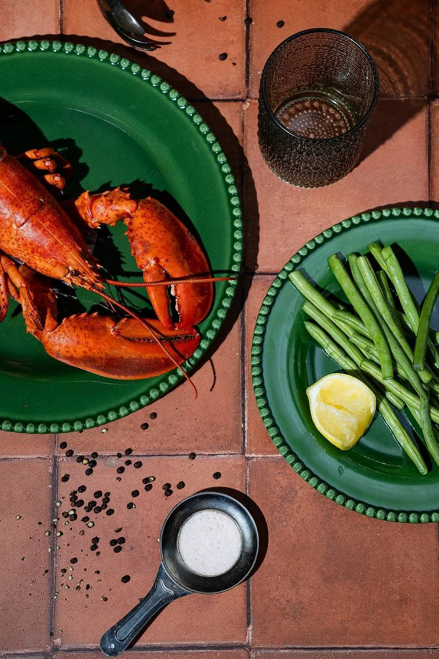 Lobster on a green plate with green beans, lemon wedge, black pepper, and salt, on a tiled table with a glass of water.