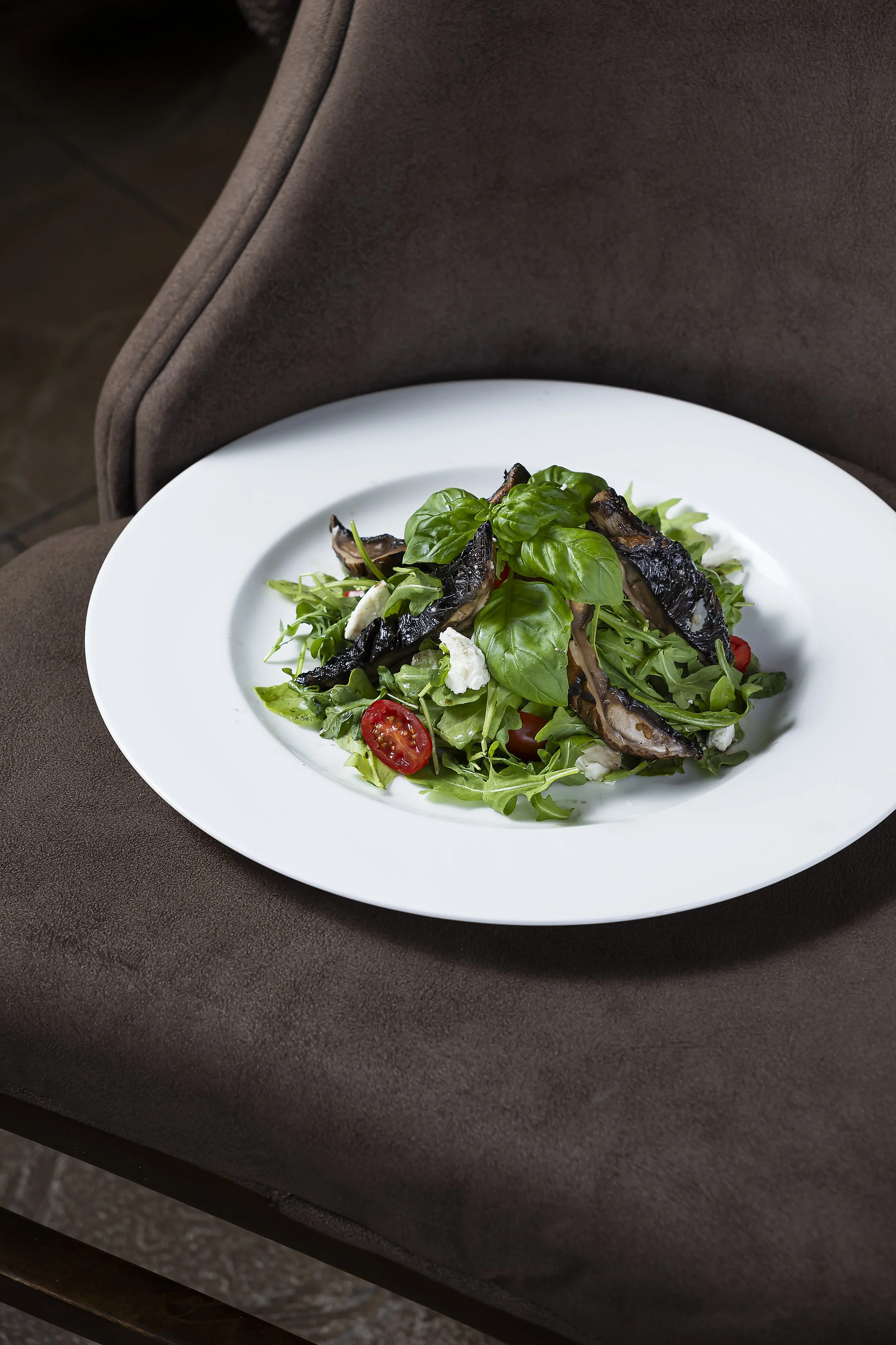 A white plate of fresh salad featuring arugula, basil leaves, cherry tomatoes, mozzarella cheese, and slices of grilled eggplant, placed on a brown table with a cushioned chair in the background.