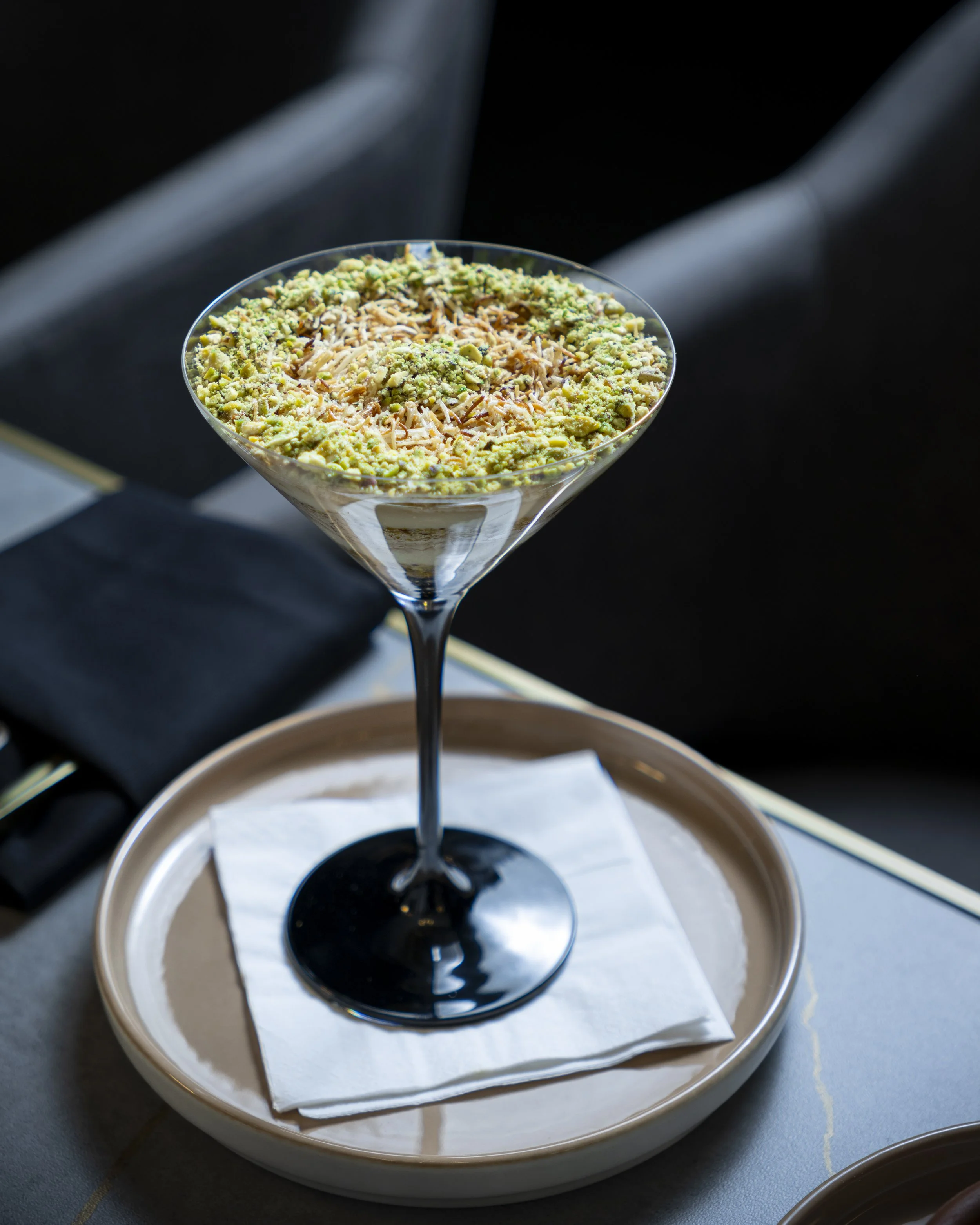 A martini glass filled with chopped pistachios and shredded cheese, placed on a paper towel on a round tray, with a black and gray background.