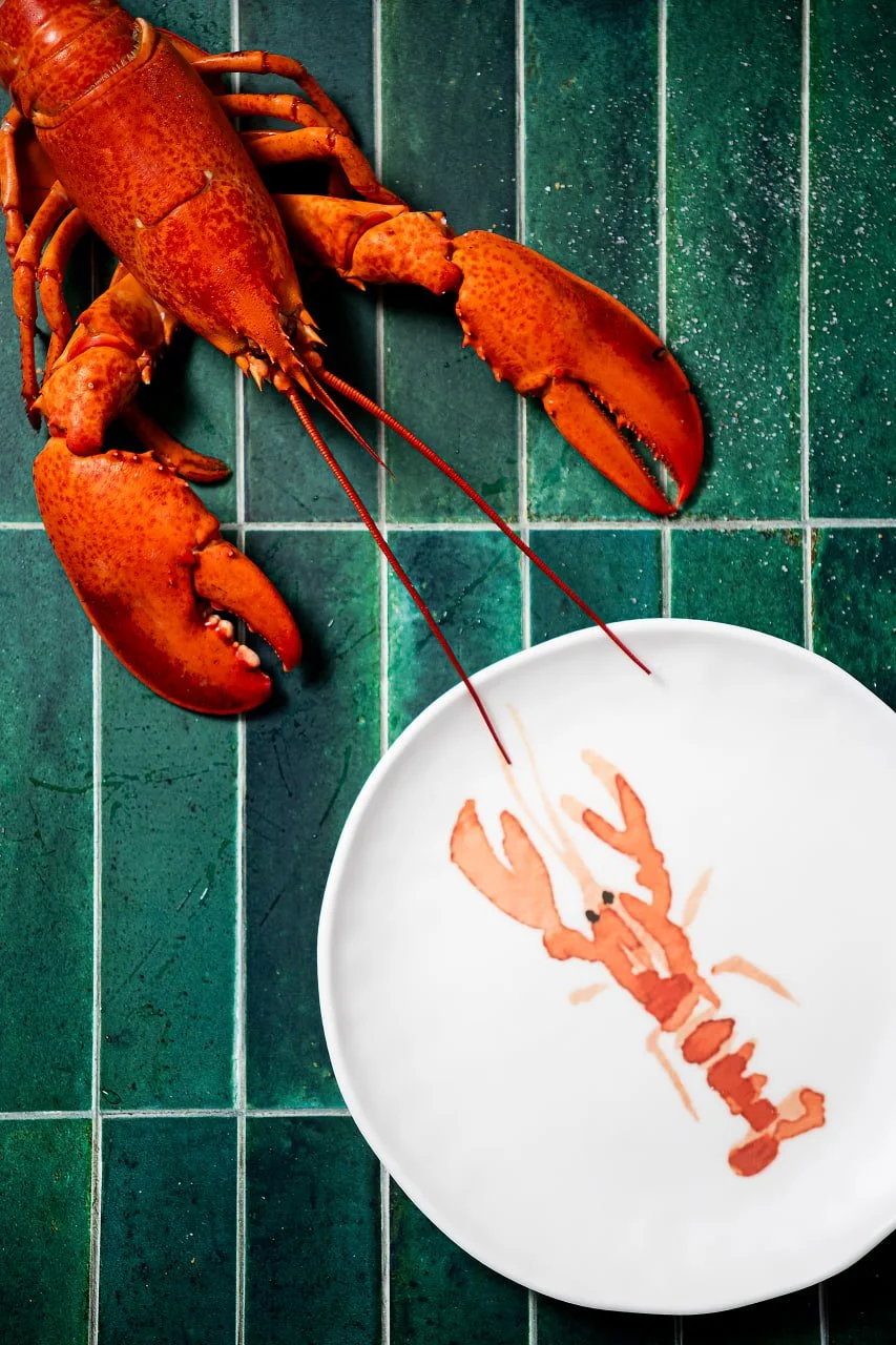 A cooked lobster on a green tiled surface next to a white plate with a lobster graphic on it.