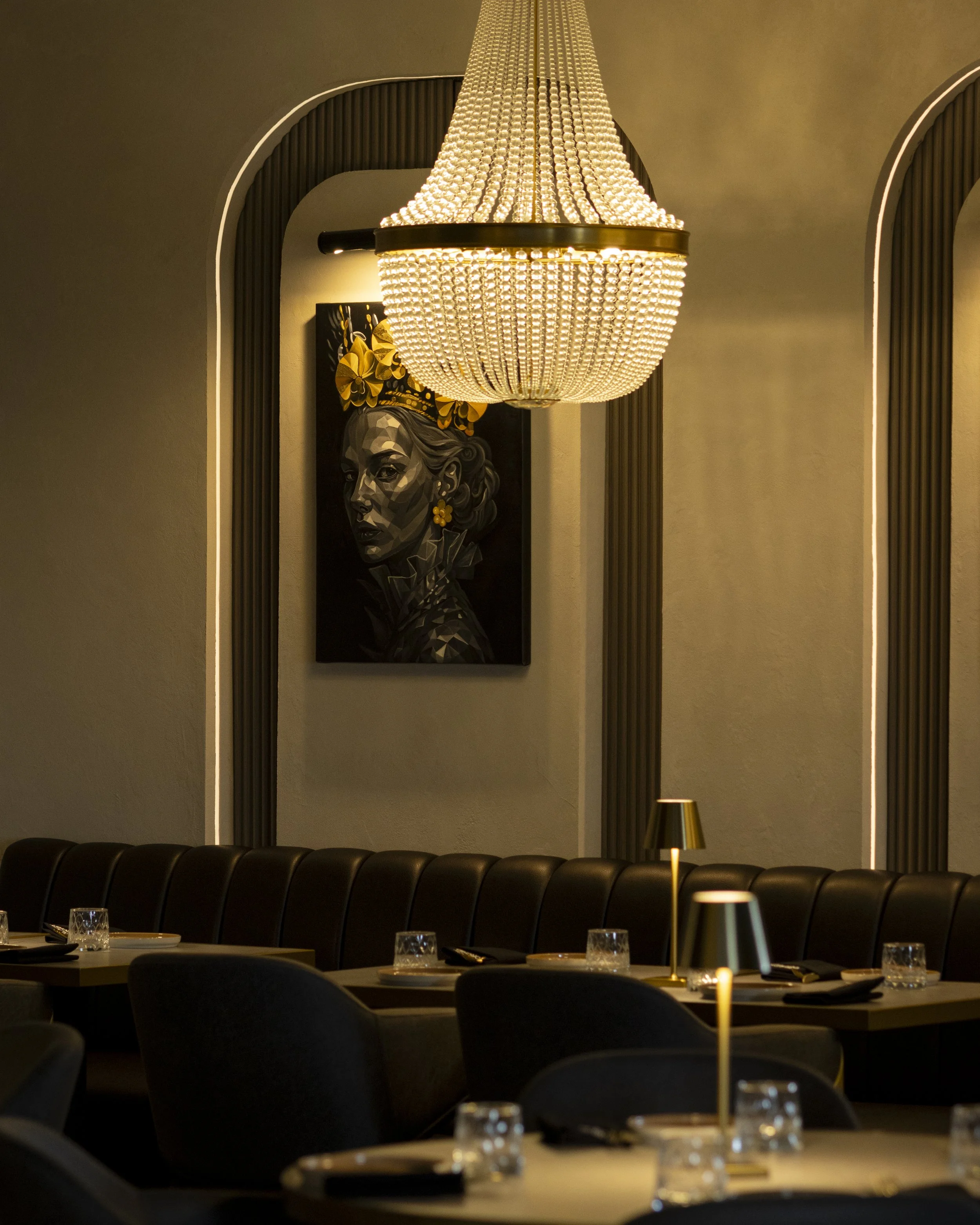 A restaurant dining area with a large chandelier hanging from the ceiling. There are tables set with glassware and chopsticks, and a black leather banquette along the wall. A framed portrait of a woman's face with a yellow floral headdress is on the 