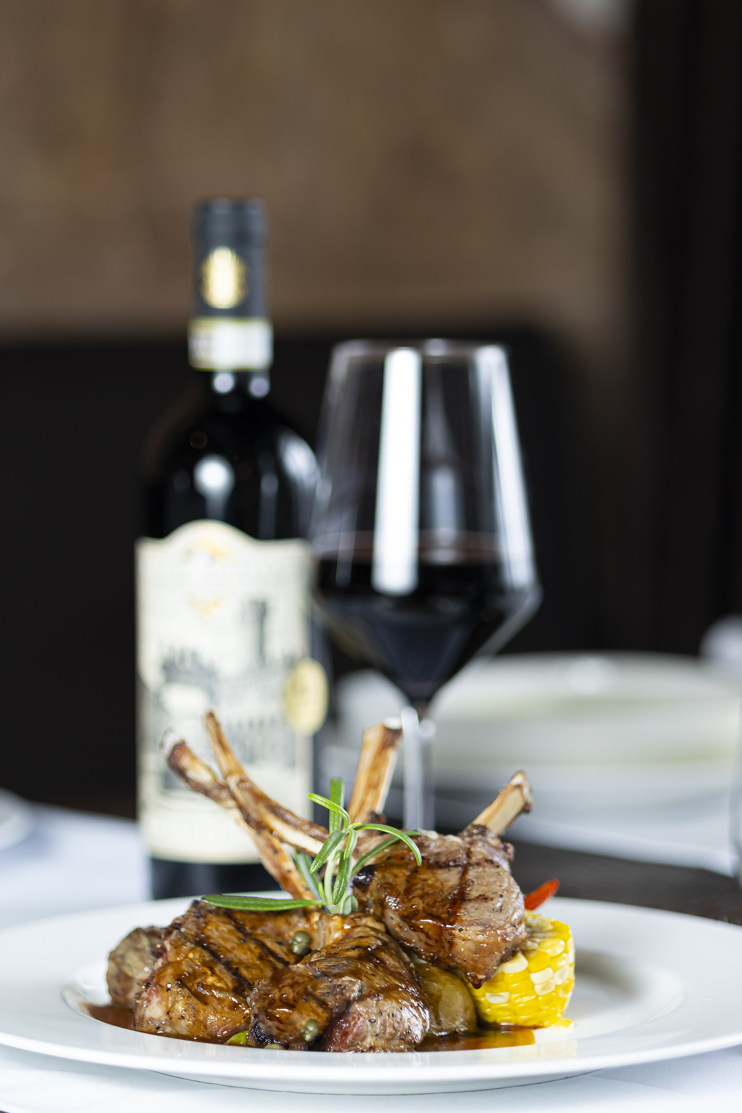 A plate of cooked lamb chops garnished with rosemary, served with a piece of corn on the cob, with a bottle of wine, a glass of red wine, and a blurred background.