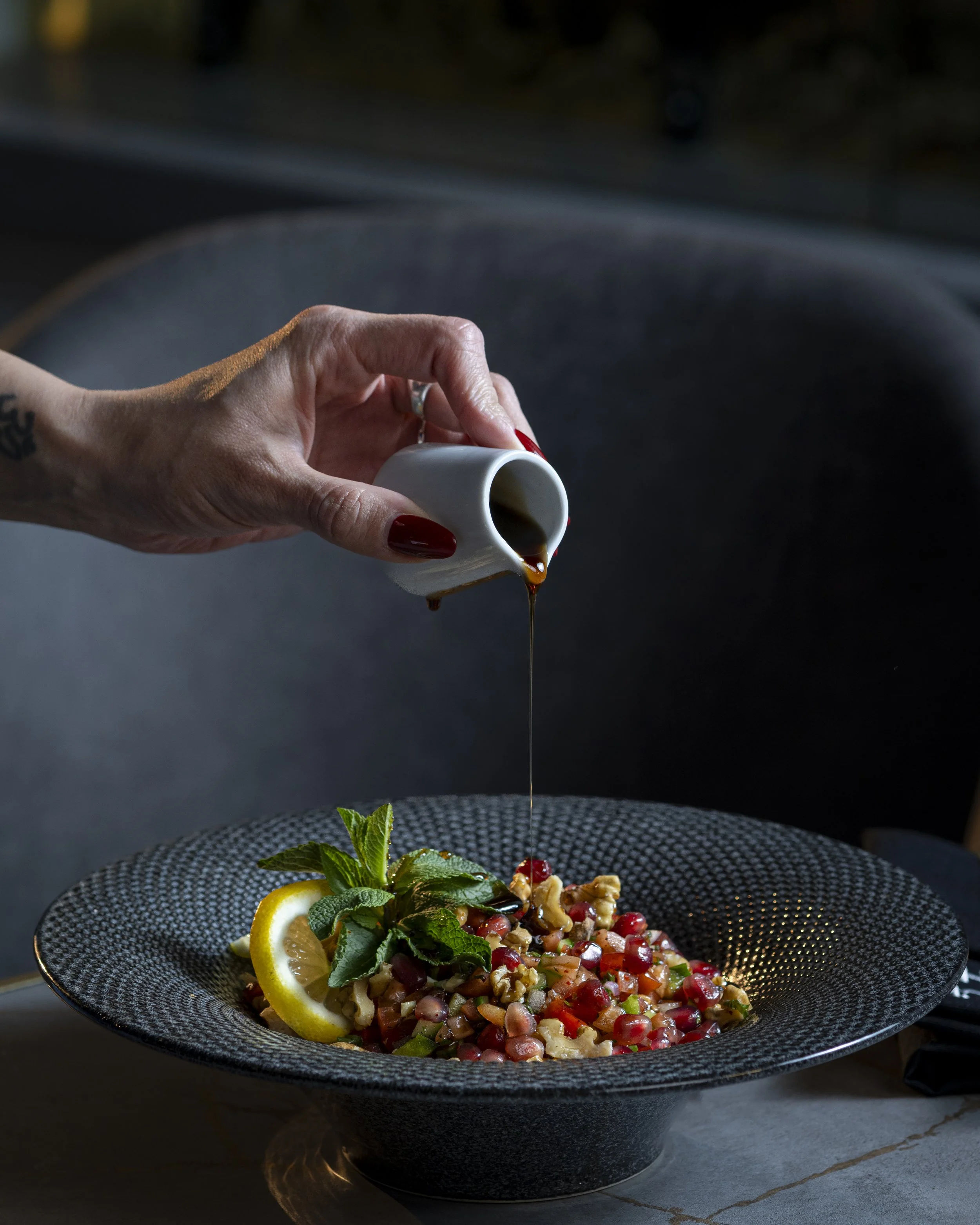 A hand pouring dark balsamic vinegar over a colorful pomegranate and veggie salad garnished with lemon slices, mint, and walnuts in a black textured bowl.