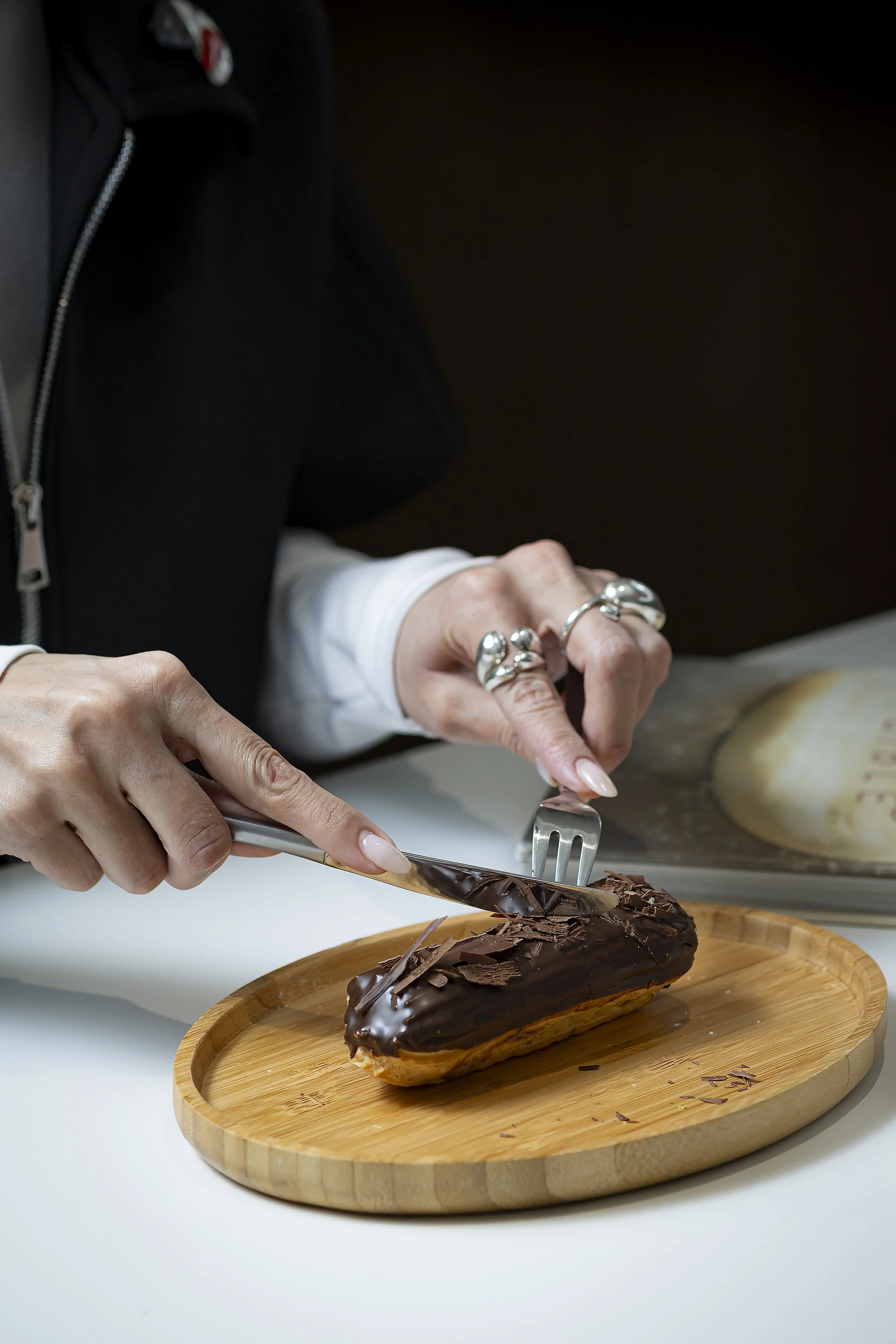 A person is cutting a chocolate éclair with a knife on a wooden plate. The person is wearing jewelry, including rings and a bracelet, and has a black jacket and white sleeve visible. The éclair is covered in chocolate and placed on a round wooden pla