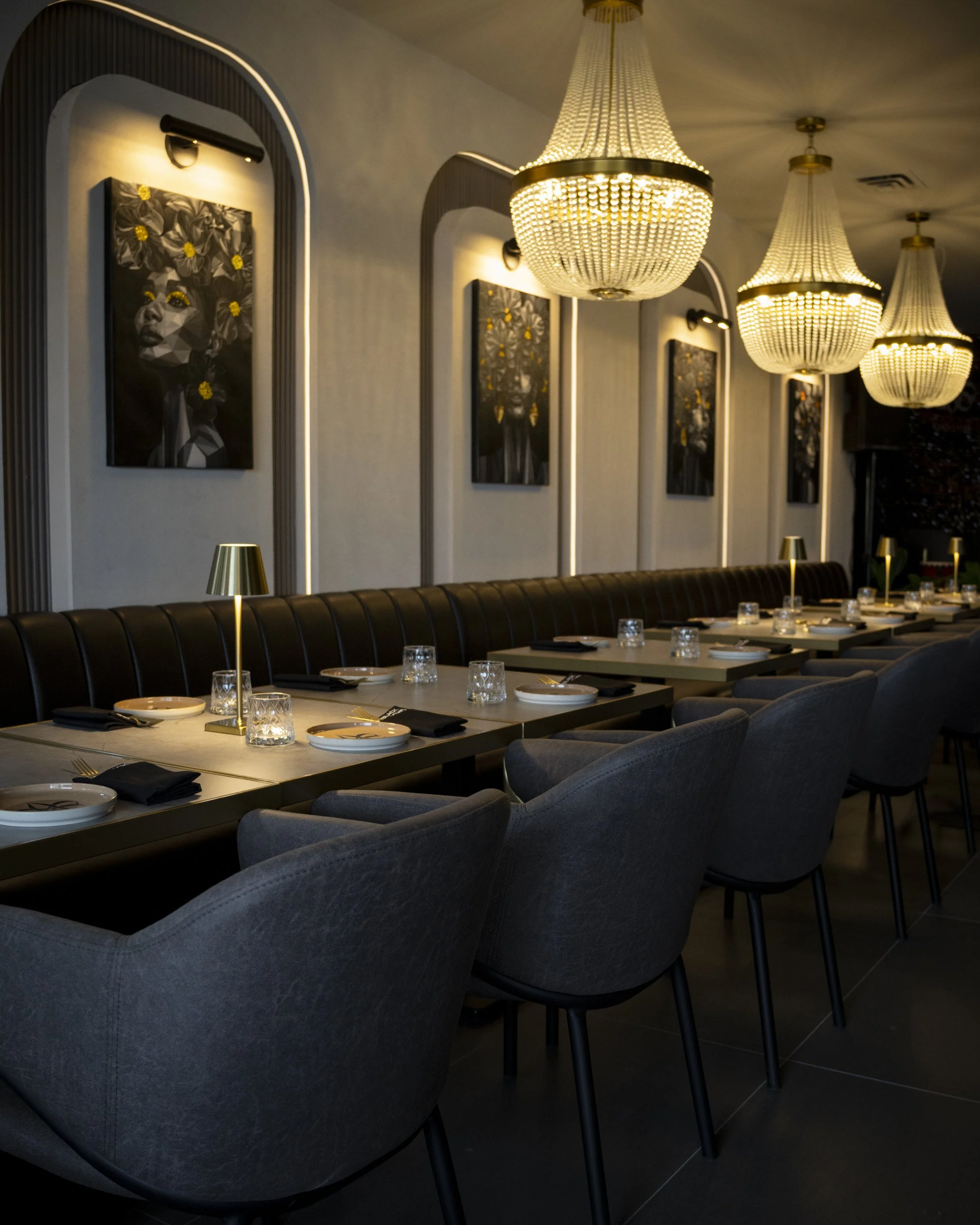 Elegant dining area with a long table set with plates, glasses, and black napkins. Modern gray upholstered chairs surround the table. The room features large, ornate chandeliers hanging from the ceiling, and artworks of painted faces with floral acce