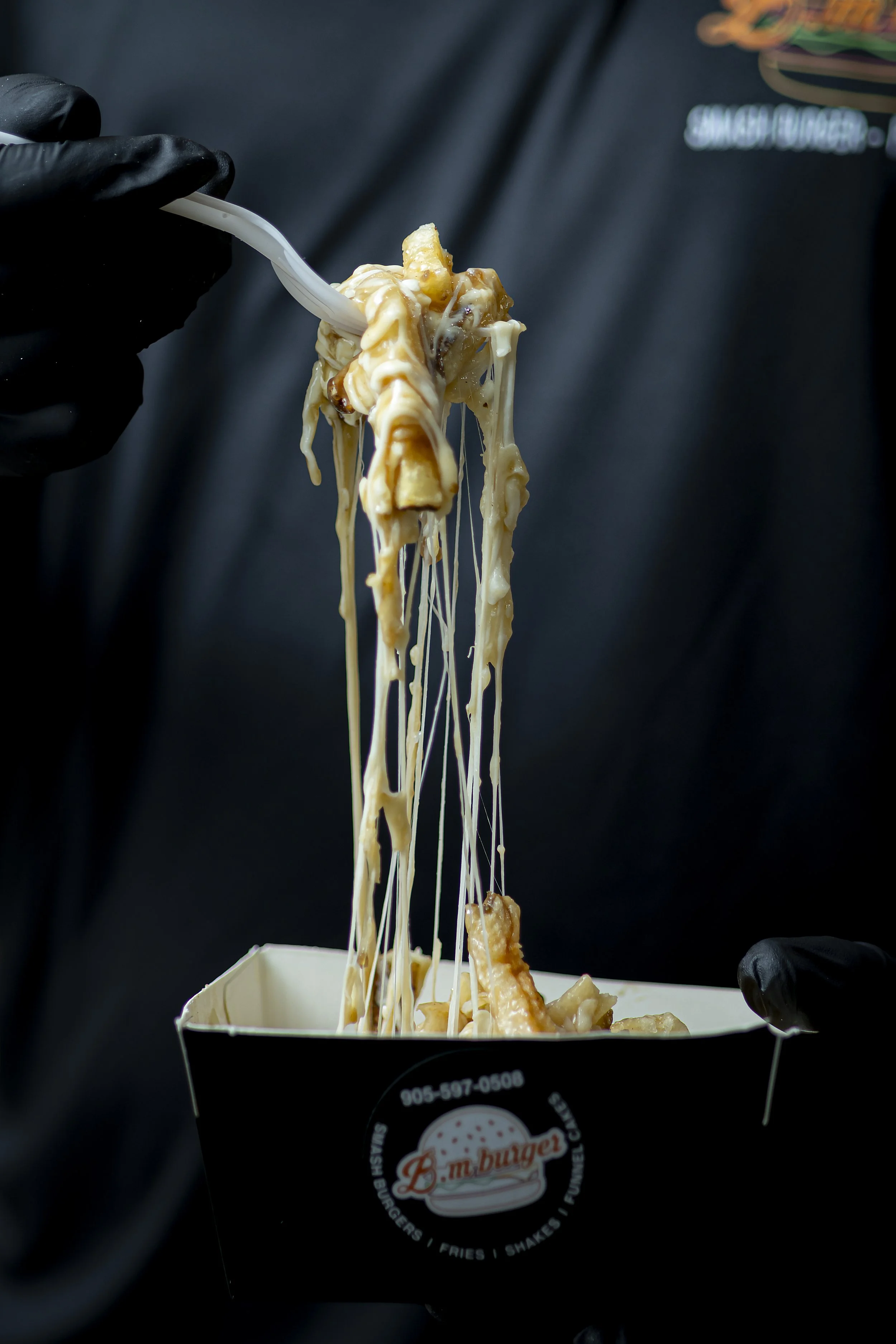 A person wearing black gloves holding a fork with cheesy fries from B.M. Burger in a takeout container.