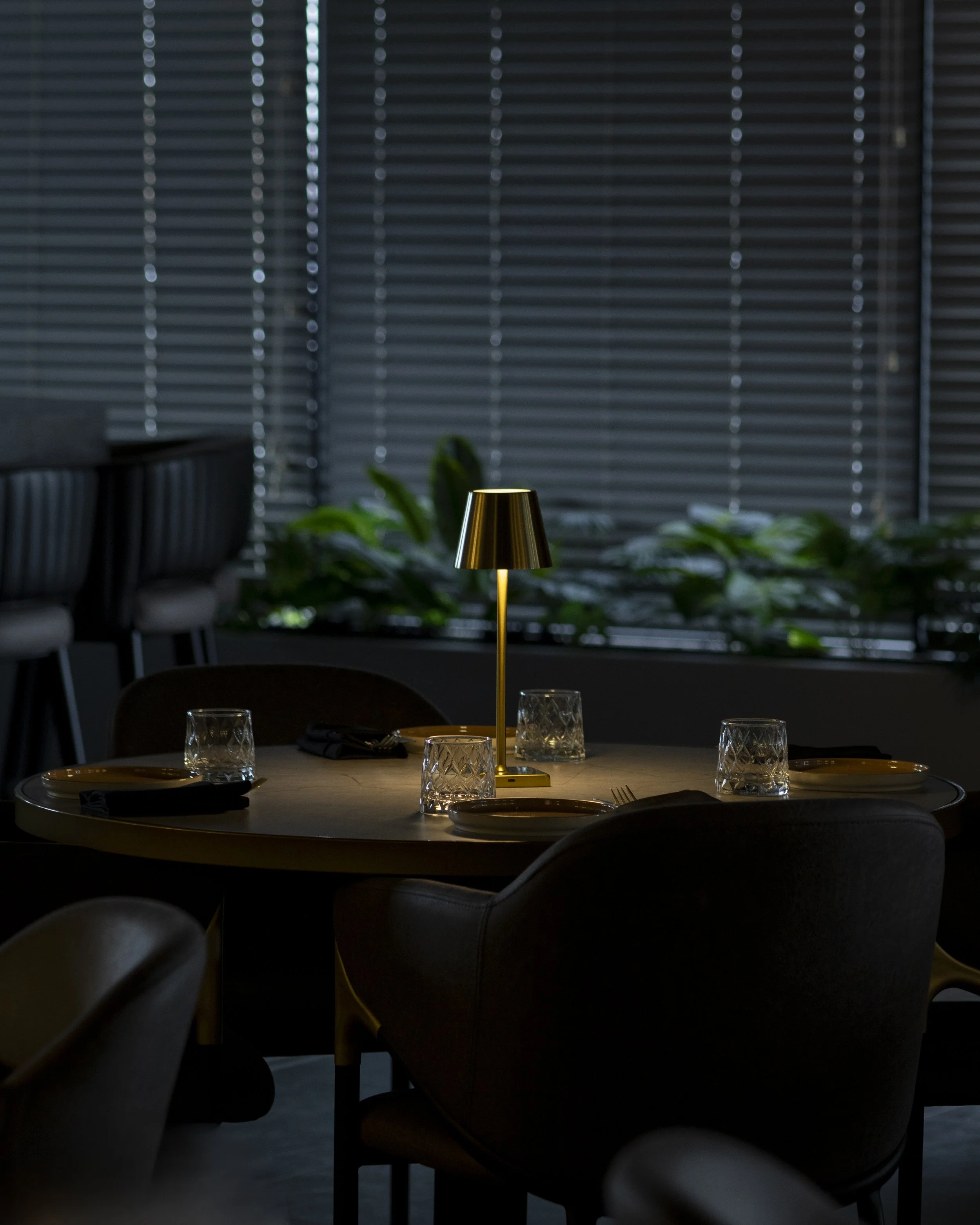 A dimly lit dining room table with a gold table lamp in the center, surrounded by glasses and plates, with closed horizontal blinds and green plants in the background.