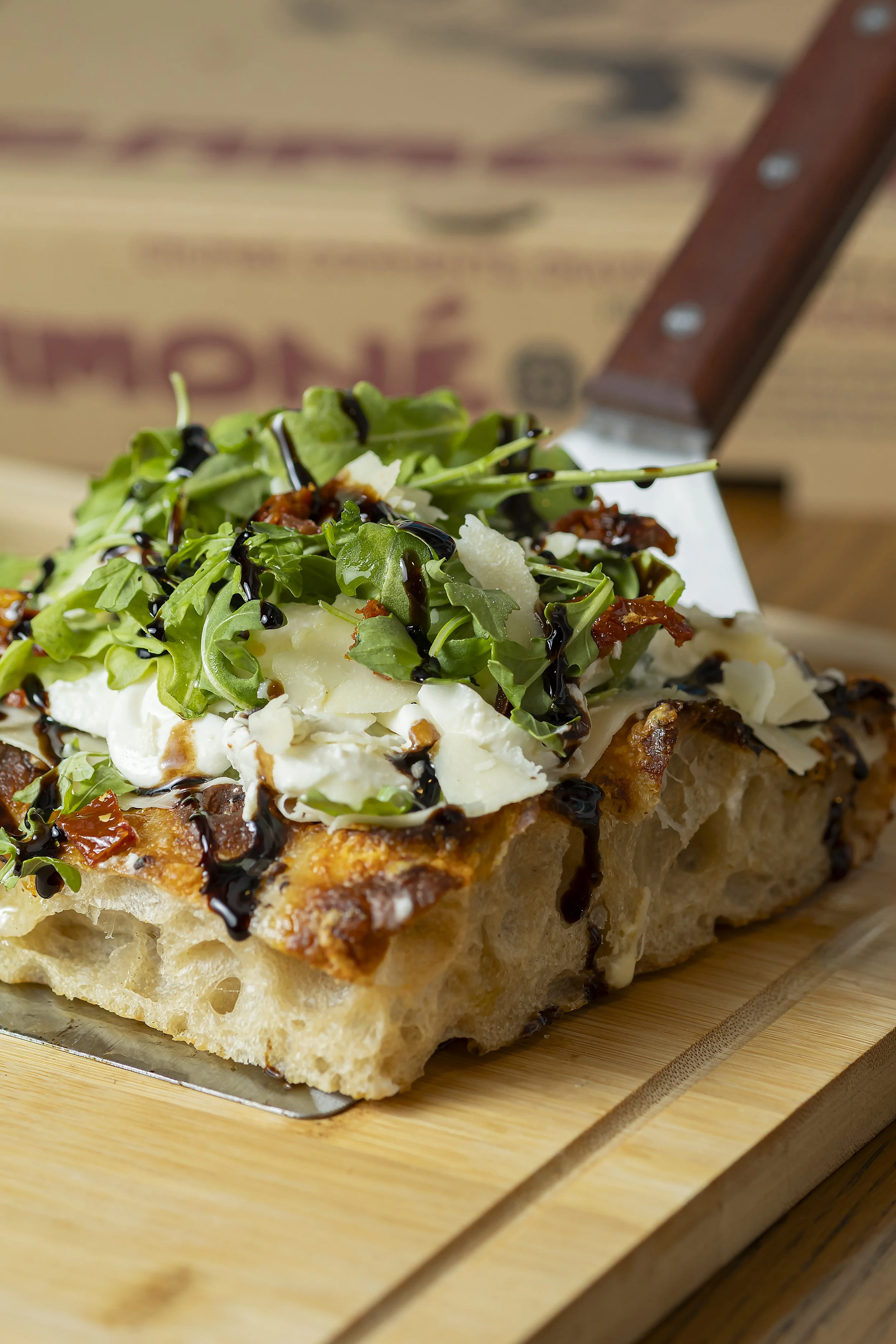 Close-up of a slice of pizza with arugula, shaved cheese, sun-dried tomatoes, balsamic glaze, and a crispy crust on a wooden board.